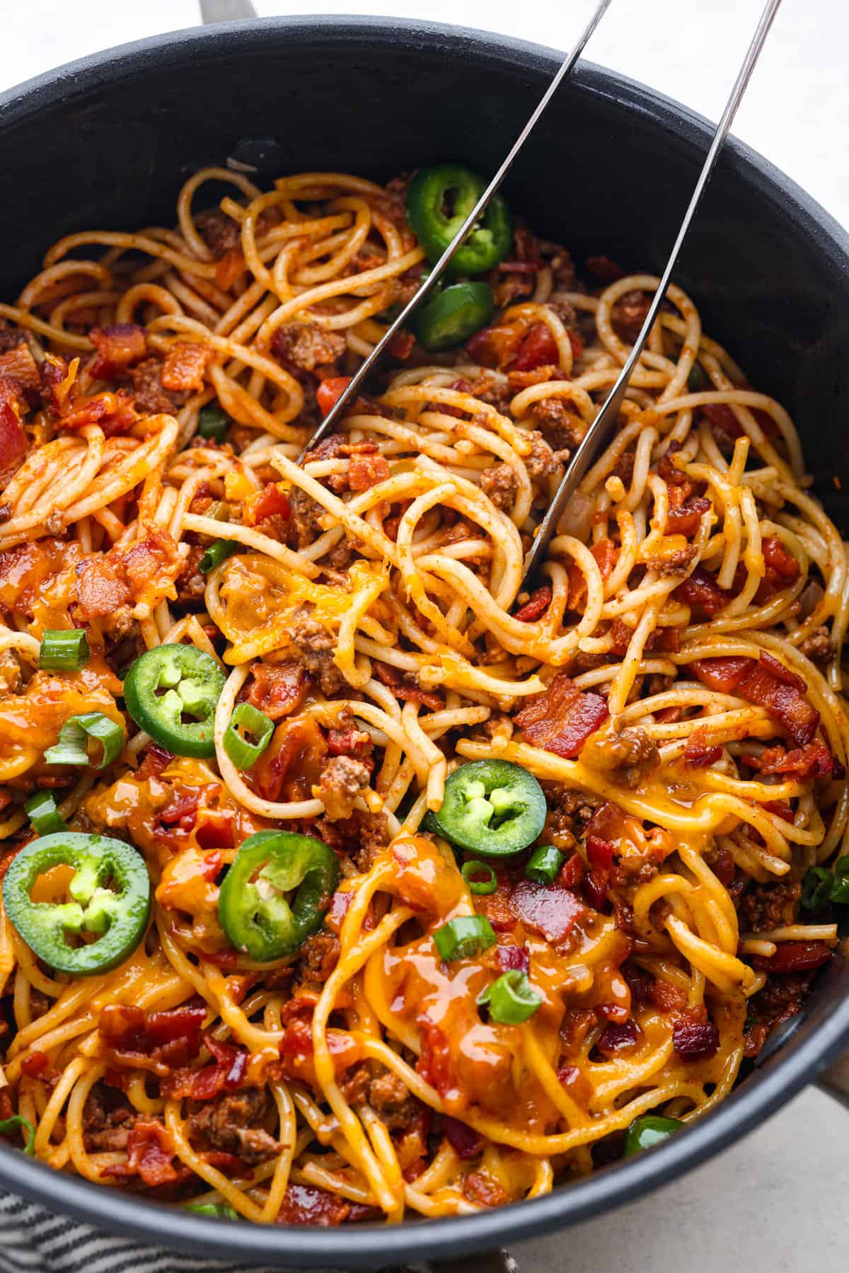 A skillet filled with spaghetti mixed with ground meat, tomato sauce, melted cheese, and topped with slices of fresh green jalapeño peppers is perfect for busy families. Two forks are inserted into the pasta
