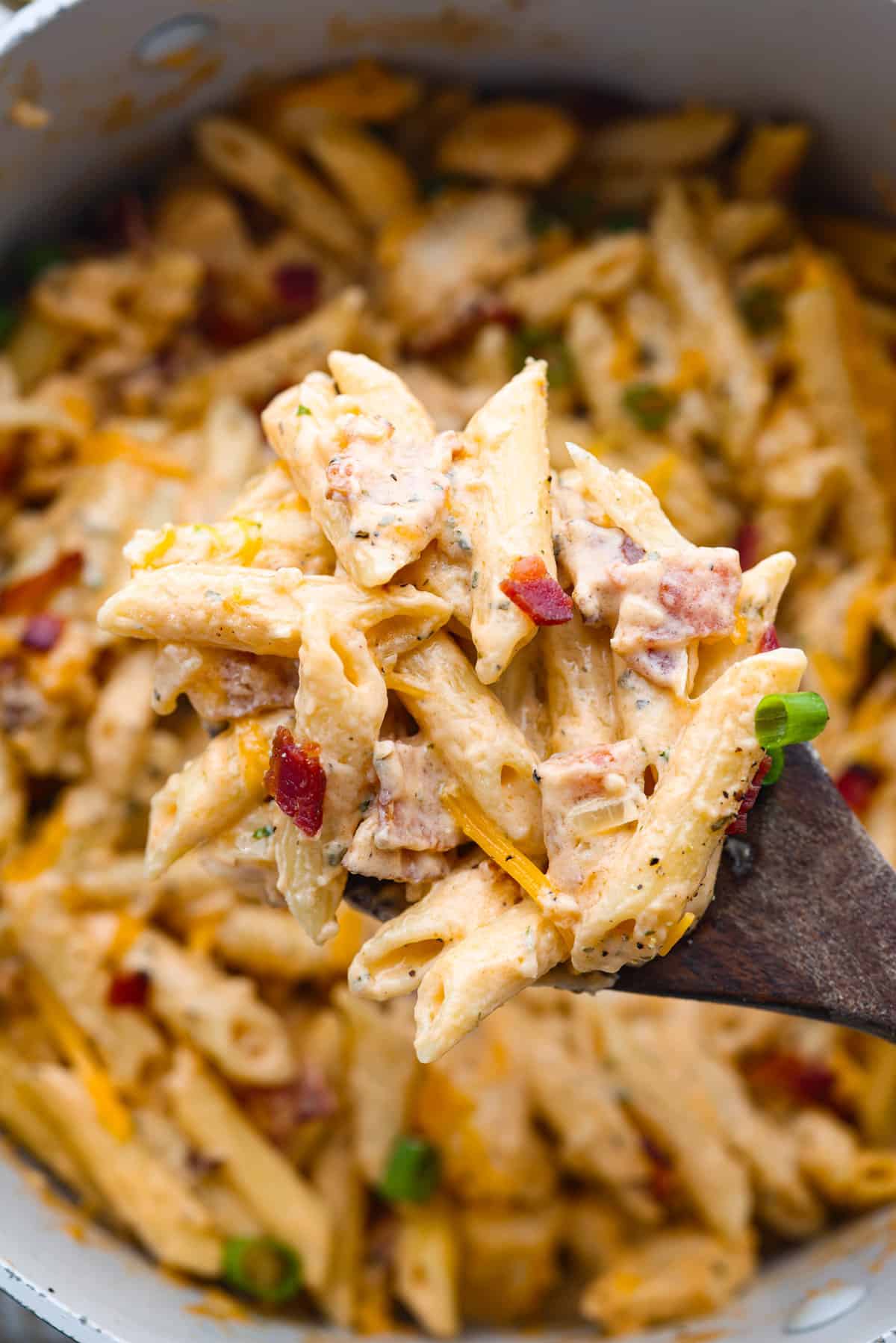 A close-up view of creamy chicken pasta with red peppers and green onions in a pot, perfect for easy dinner ideas, with a wooden spoon lifting some of the dish.