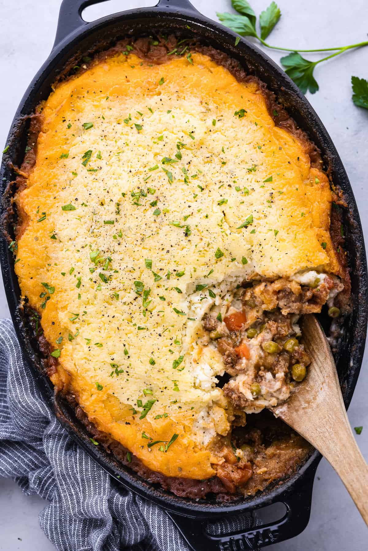 A shepherd's pie in a cast iron skillet, topped with golden-brown mashed potatoes, sprinkled with herbs. Perfect for busy families, a wooden spoon scoops into the pie, revealing the meat