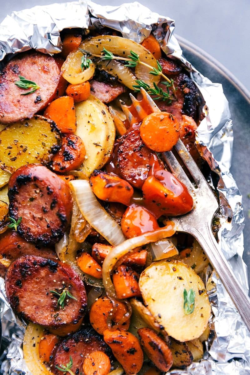 Grilled sausage and mixed vegetables including sliced potatoes, carrots, and onions, garnished with parsley in a foil packet. An easy dinner idea perfect for busy families, served with a fork.