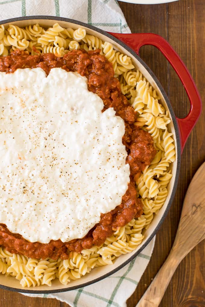A pot of cooked fusilli pasta topped with a thick tomato meat sauce and a generous dollop of ricotta cheese, garnished with black pepper, served in a red enamel pot. Ideal for busy