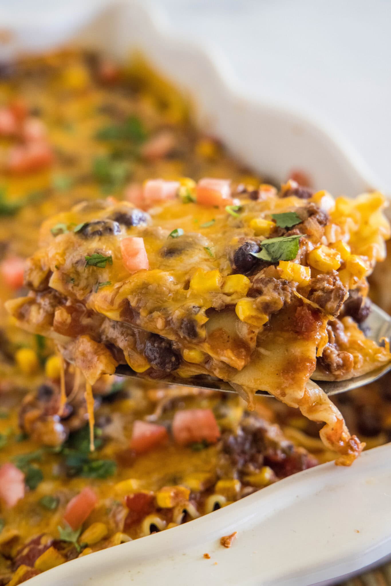 An image showcasing a spoon lifting a piece of mexican lasagna from a white dish topped with fresh parsley, 