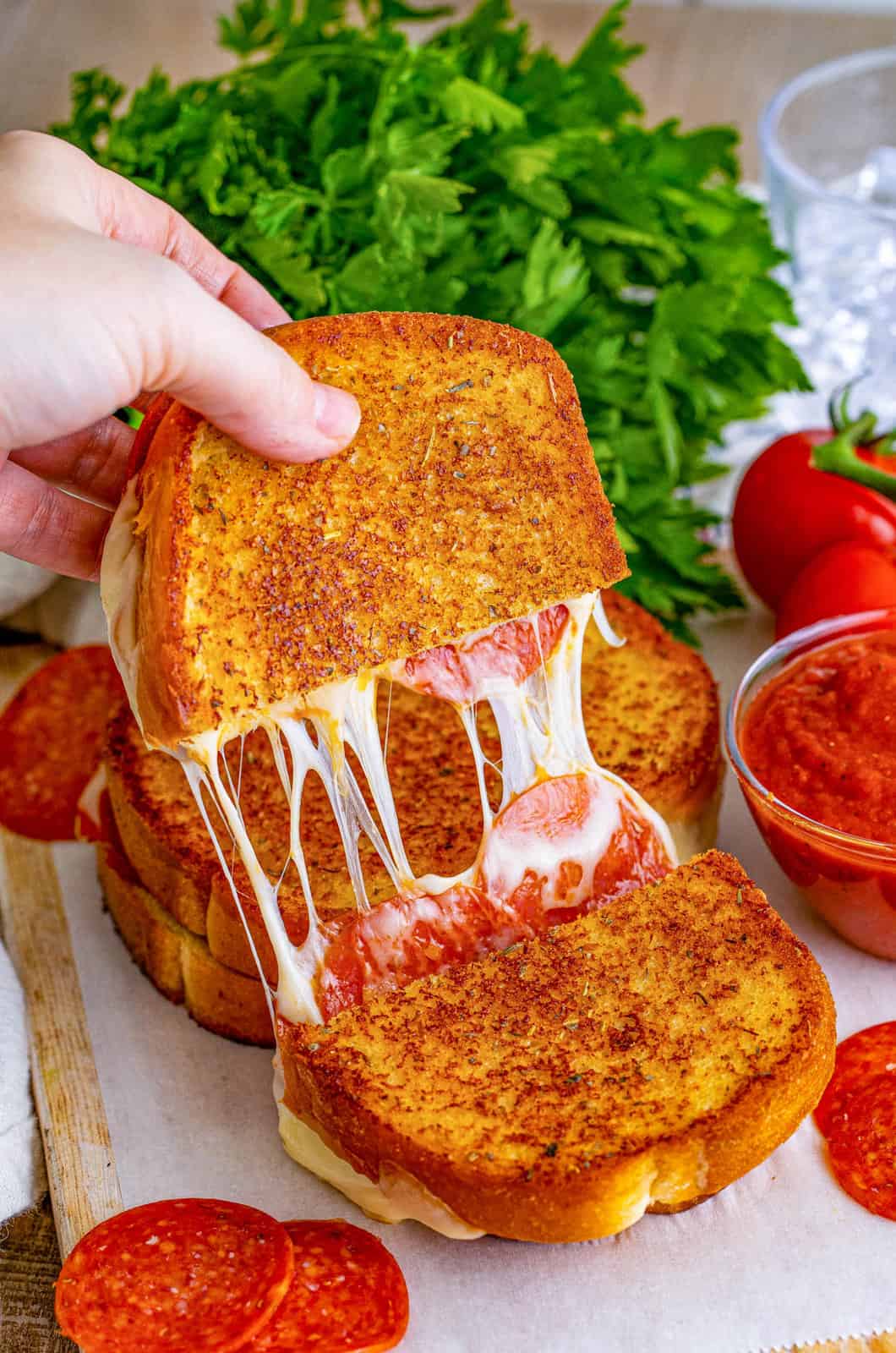A person's hand holding a gooey cheese pull from a slice of pepperoni pizza toast, perfect for easy dinner ideas, with fresh tomatoes, parsley, and more pepperoni slices visible in the background