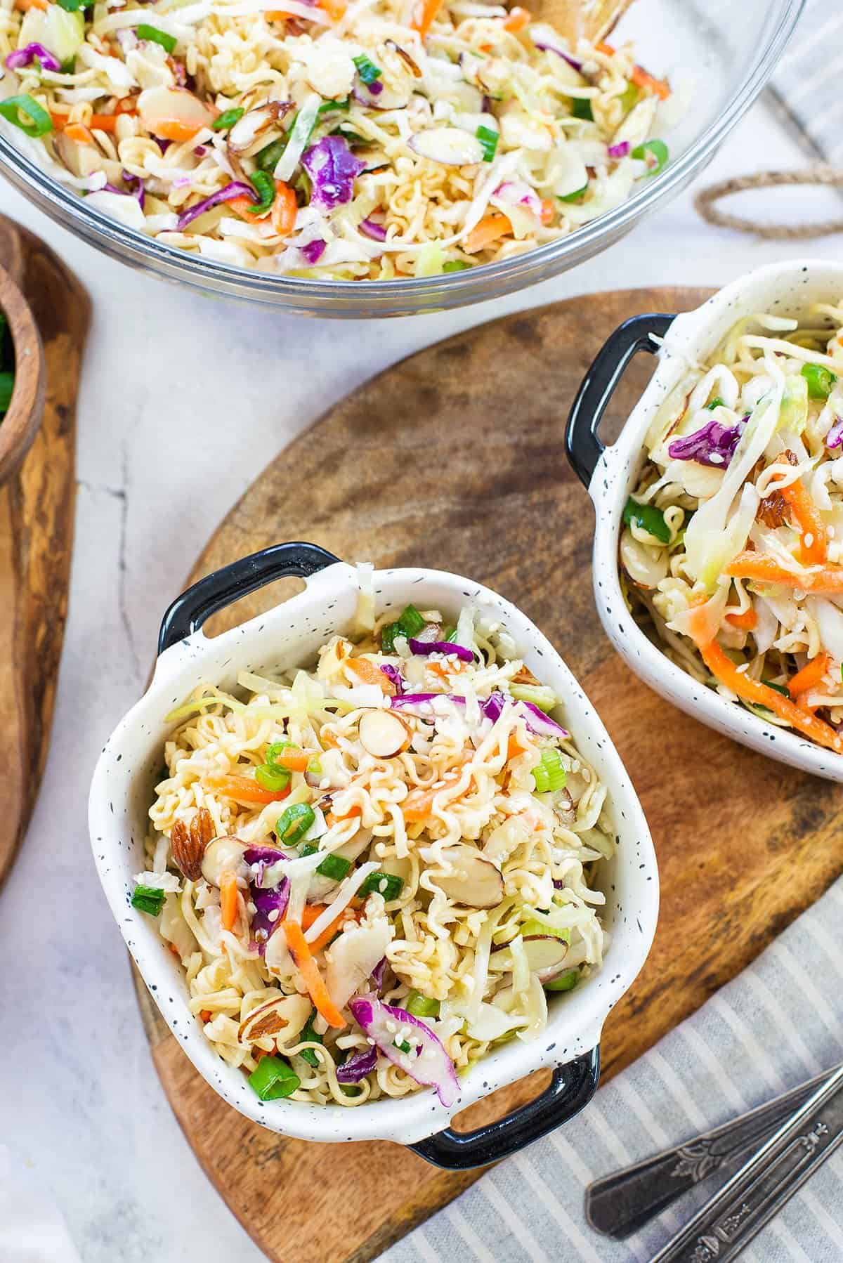 Two white bowls filled with colorful Asian-inspired ramen noodle salad made of shredded cabbage, carrots, scallions, and ramen noodles, garnished with sesame seeds, on a white marble surface