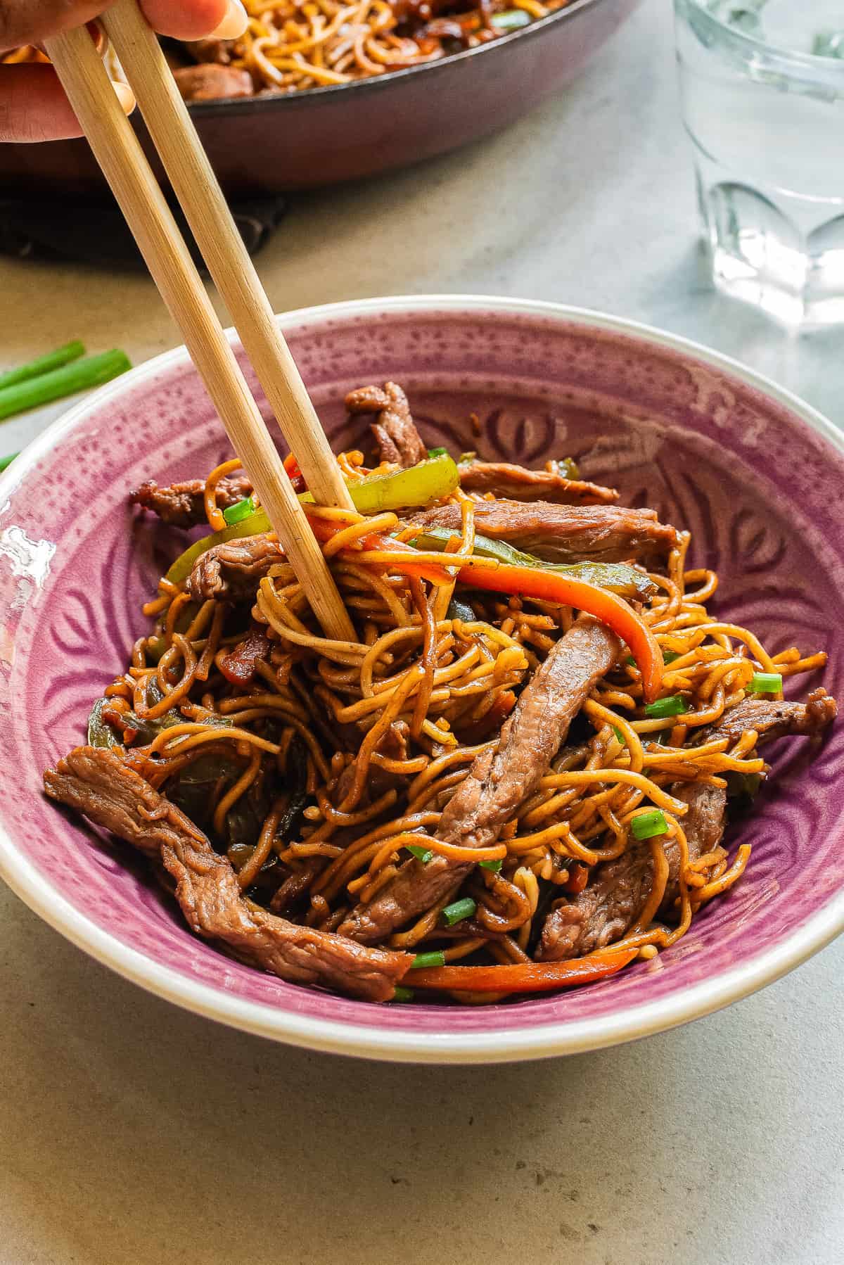 A bowl of stir-fry noodles with beef and sliced vegetables, being eaten with chopsticks. The noodles appear savory and are garnished with green onions, served in a decorative pink bowl. This dish