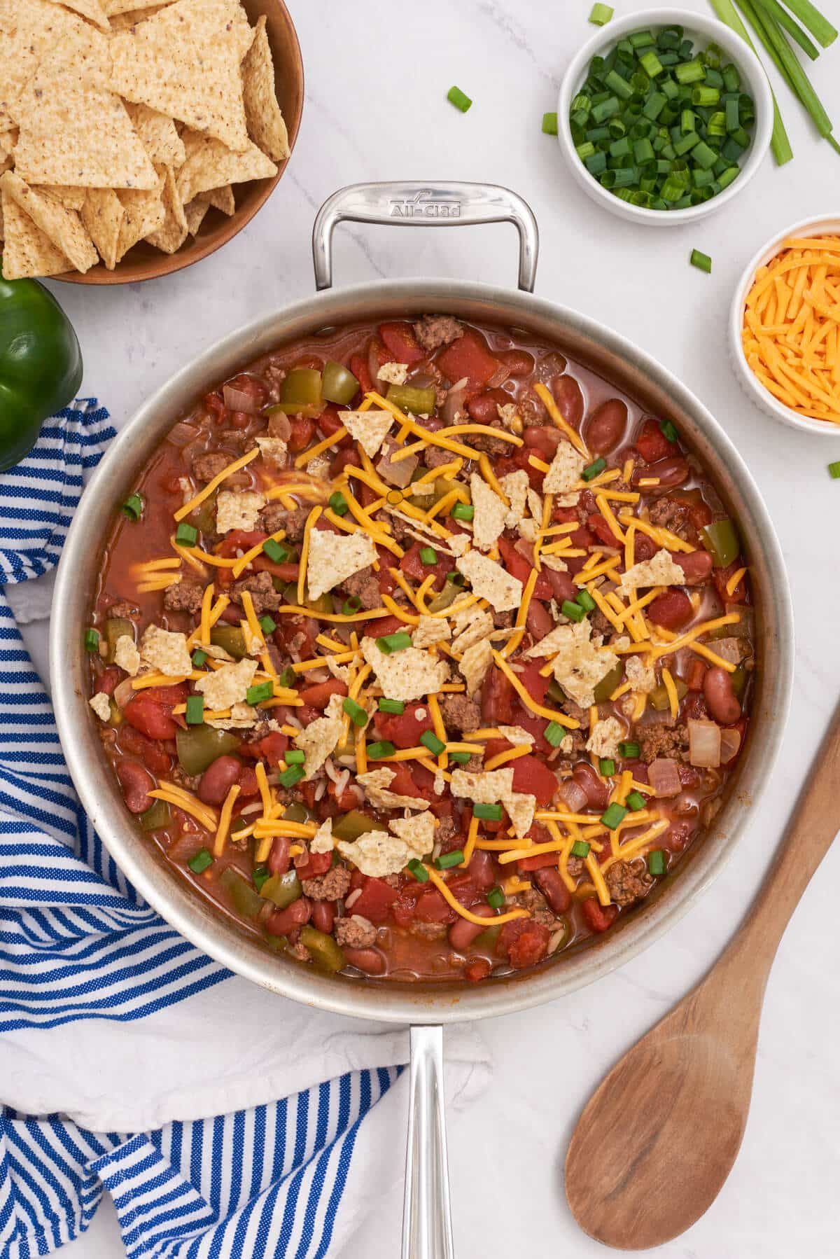 A stainless-steel skillet filled with chili topped with shredded cheese, crushed tortilla chips, and diced green onions—perfect for busy families—alongside a bowl of tortilla chips and a wooden spoon