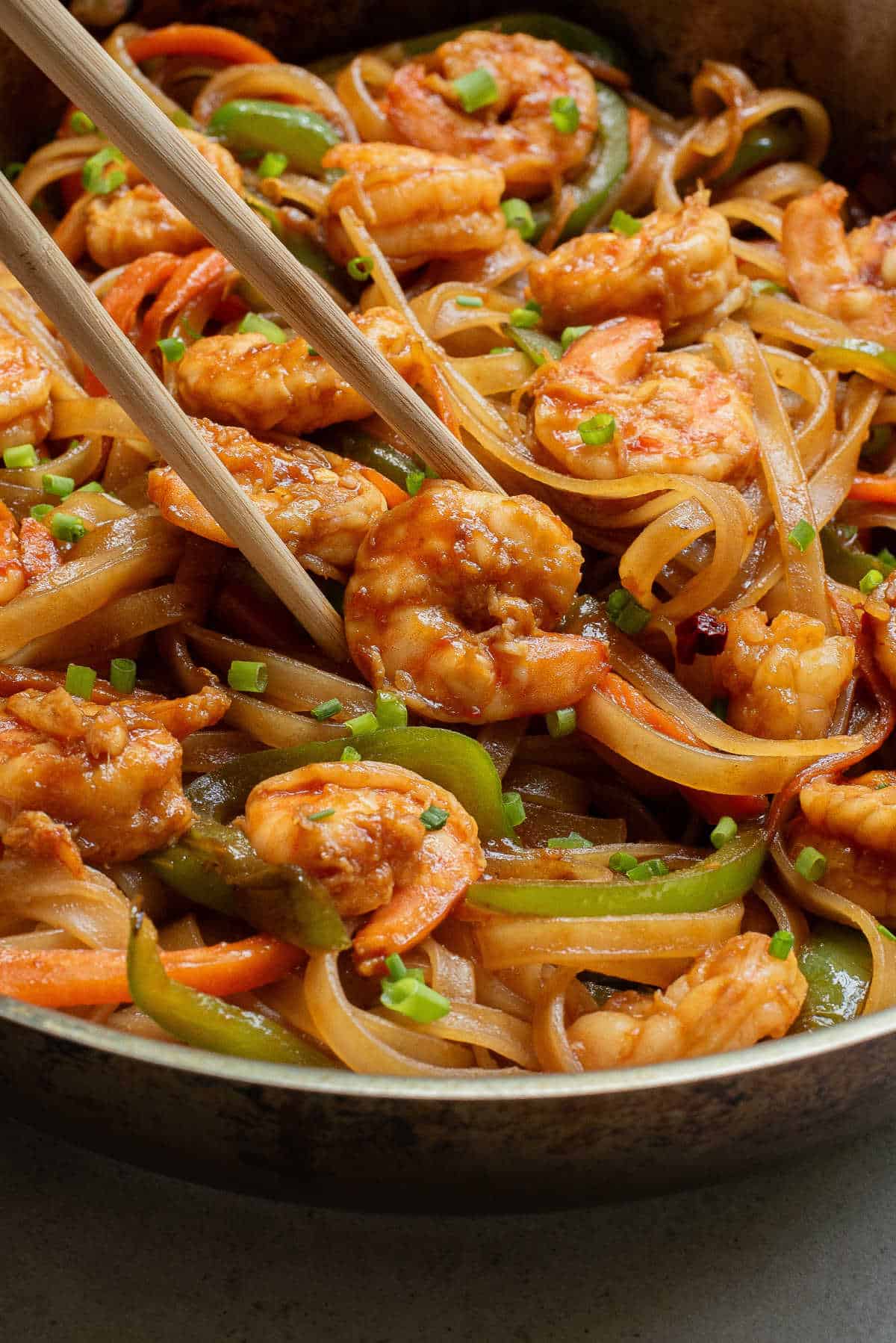 Close-up image of an easy stir-fried shrimp and vegetable dinner with noodles, vibrant green bell peppers, and carrots, served in a dark bowl with chopsticks.