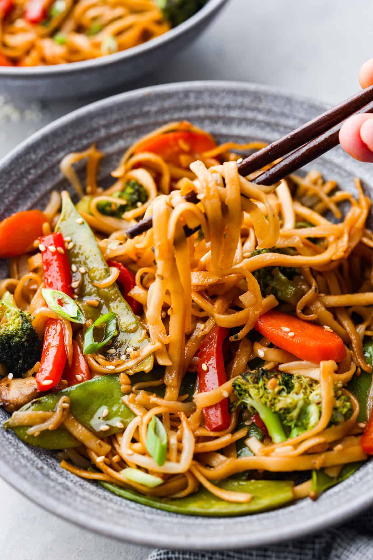 A person uses chopsticks to lift stir-fried noodles and vegetables from a dark ceramic bowl, which includes red peppers, carrots, and broccoli—an easy dinner idea for busy families.
