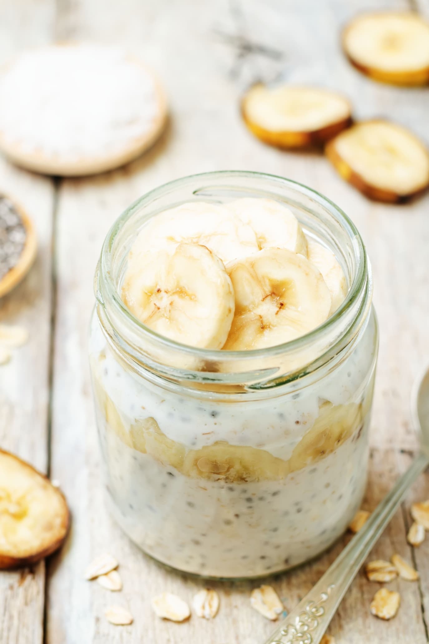 A jar of overnight oats with banana and chia seeds, set on a rustic wooden table. The foreground shows a clear glass jar layered with creamy oats speckled with tiny, black chia seeds, topped with several round slices of ripe banana. In the background, there's a blurred view of a whole banana cut in half and some scattered oats. The golden tones of the wood and the soft, natural light lend a cozy, inviting feel to this nutritious breakfast option for toddlers.