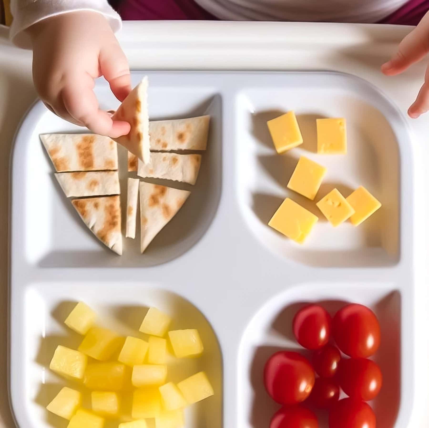 child hands reaching for a plate of toddler breakfast ideas including sliced pita bread, pineapple bites, cheese cubes and grape tomatoes