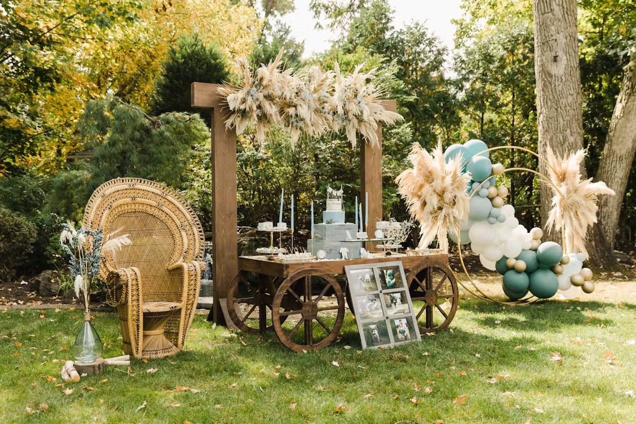 A rustic outdoor setup for a boho baby shower features a wooden cart with greenery and dried flowers, holding a three-tiered cake. To the left is a wicker peacock chair adorned with a small bouquet. To the right, a circular balloon arch incorporates pampas grass and earthy-toned balloons.