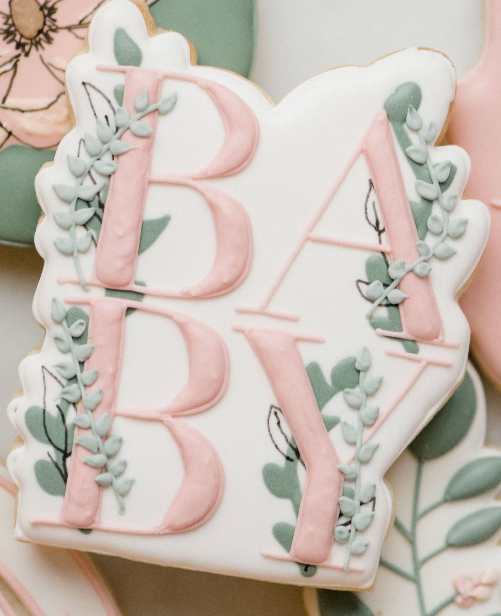 A close-up image of a creatively decorated cookie perfect for a boho baby shower. The white icing spells "BABY" in large, pink letters, with decorative green and grey leaves interwoven through the letters. Additional floral decorations are visible around the edges of the cookie.