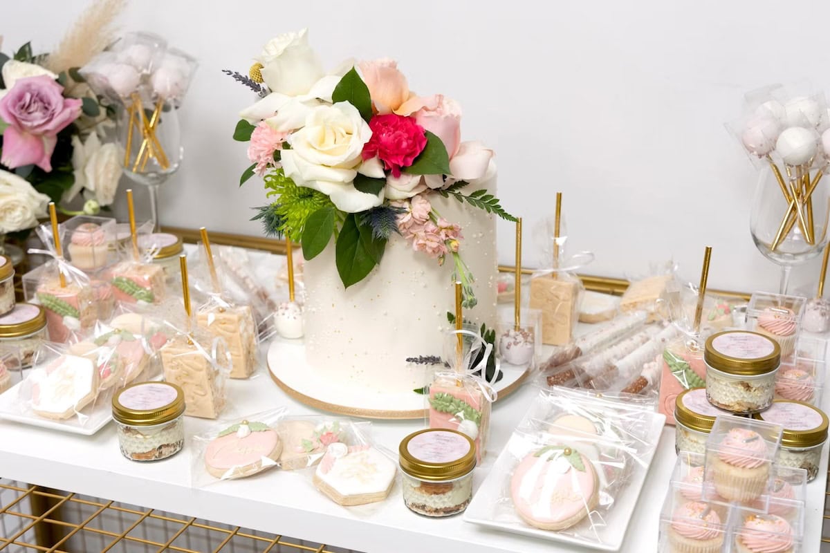 A beautifully decorated white cake with pastel flowers and greenery on top is surrounded by an array of jarred desserts, cookies, and cake pops, all elegantly packaged. The display, perfect for a boho baby shower, sits on a white table against a white backdrop with a floral arrangement beside it.