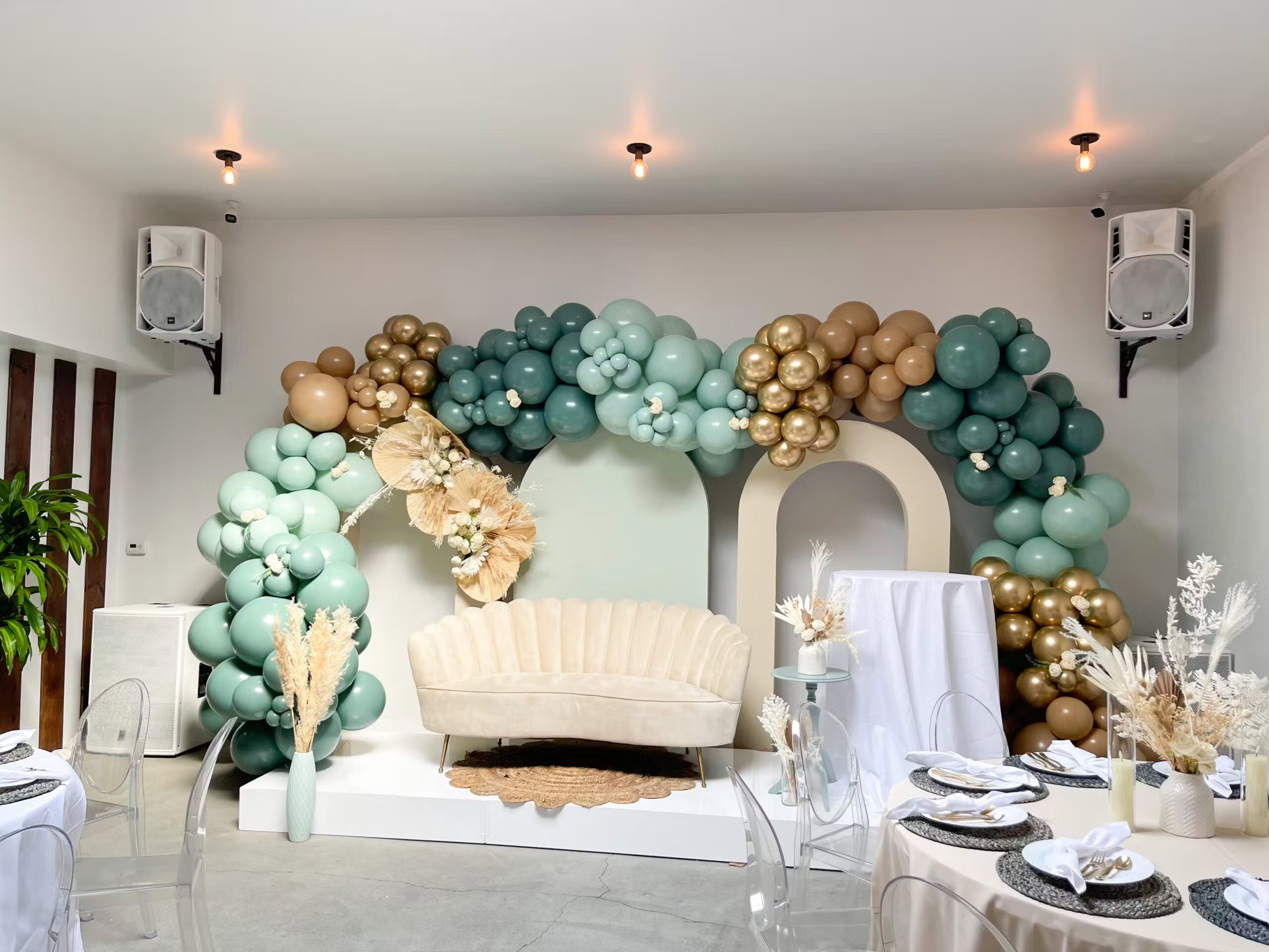 A beautifully decorated event space for a boho baby shower features a beige couch on a raised platform with a backdrop of assorted green, gold, and cream balloons. Surrounding the platform are elegantly set tables with clear chairs, white tablecloths, and dried floral centerpieces.