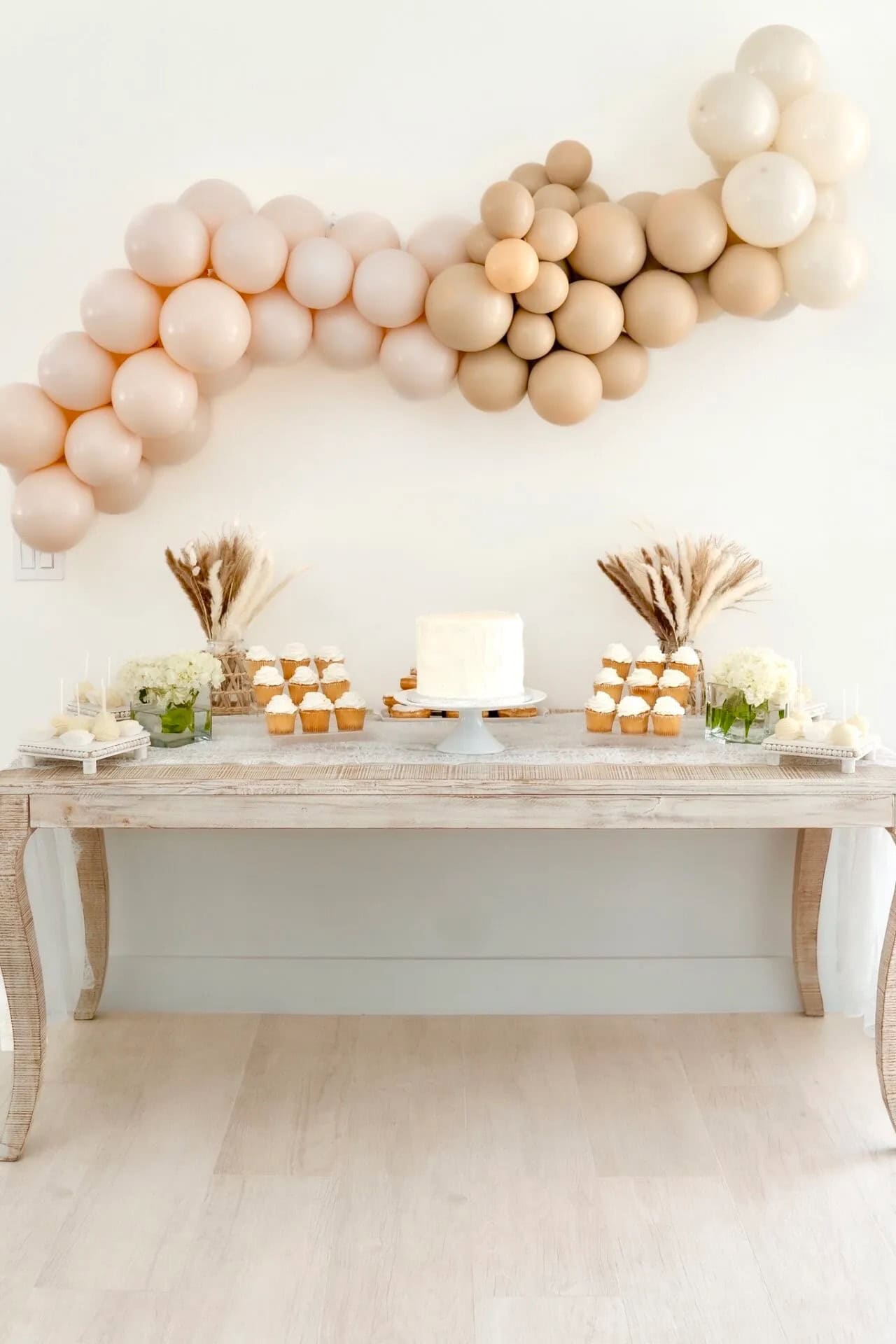 A dessert table with a rustic white wooden surface holds a central white frosted cake, cupcakes, and assorted pastries. Perfectly capturing the charm of a boho baby shower, a cloud-like beige and white balloon arrangement hangs above. Dried floral arrangements add decorative touches on either side of the table.