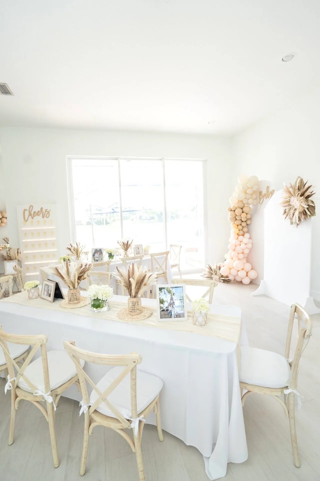 A white-themed boho baby shower decor with wooden chairs, white tables, and floral arrangements. A balloon garland is seen in the background, alongside framed pictures on tables. There's a "Cheers" sign on the left and a large sunshine-like decoration on the right wall.