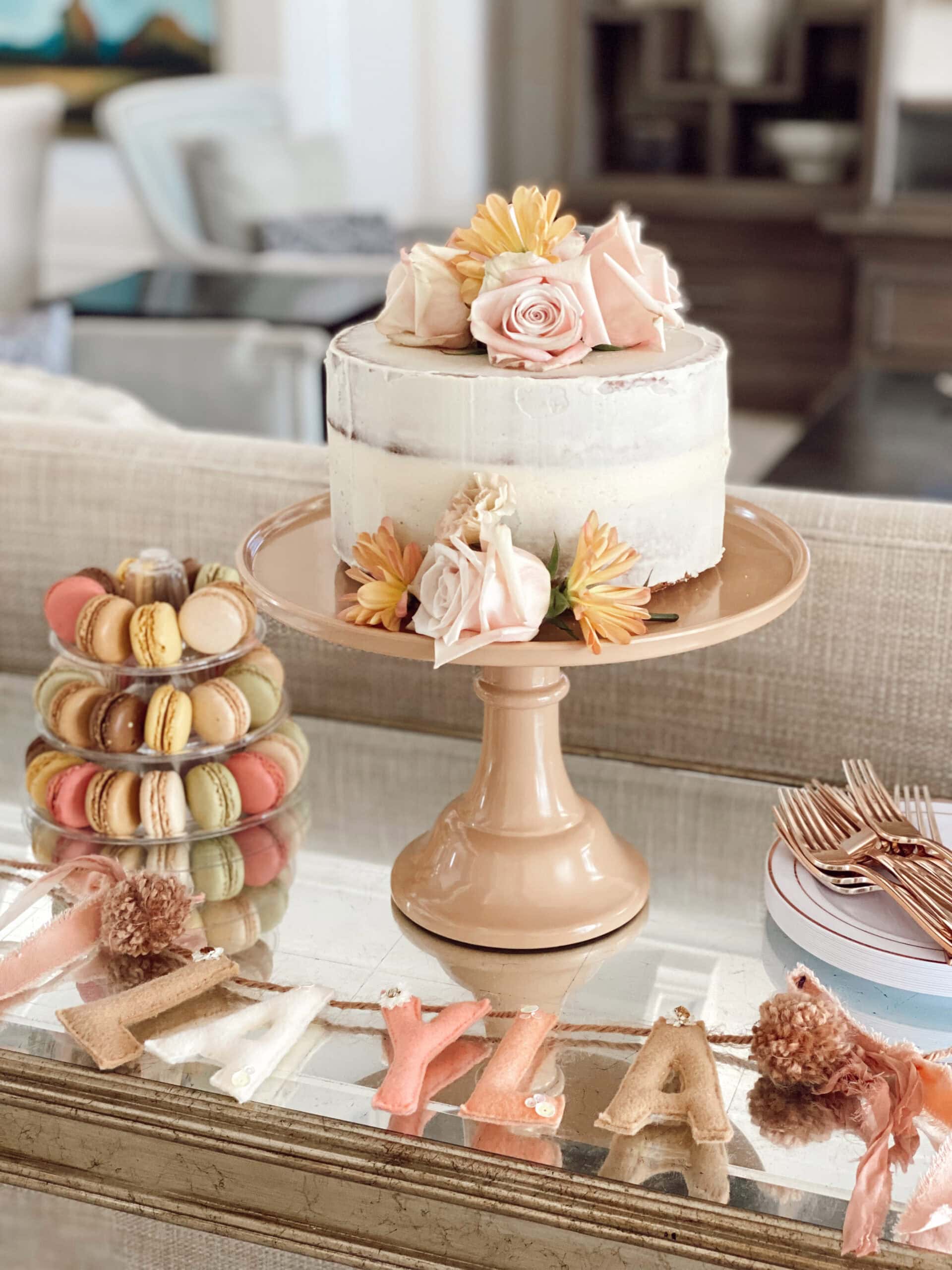 A rustic, semi-naked cake topped with pastel flowers and a single macaron sits on a peach-colored cake stand. Nearby, a tower of assorted macarons and plates with utensils are arranged on a mirrored surface. The background shows a blurred living room, perfect for a boho baby shower.