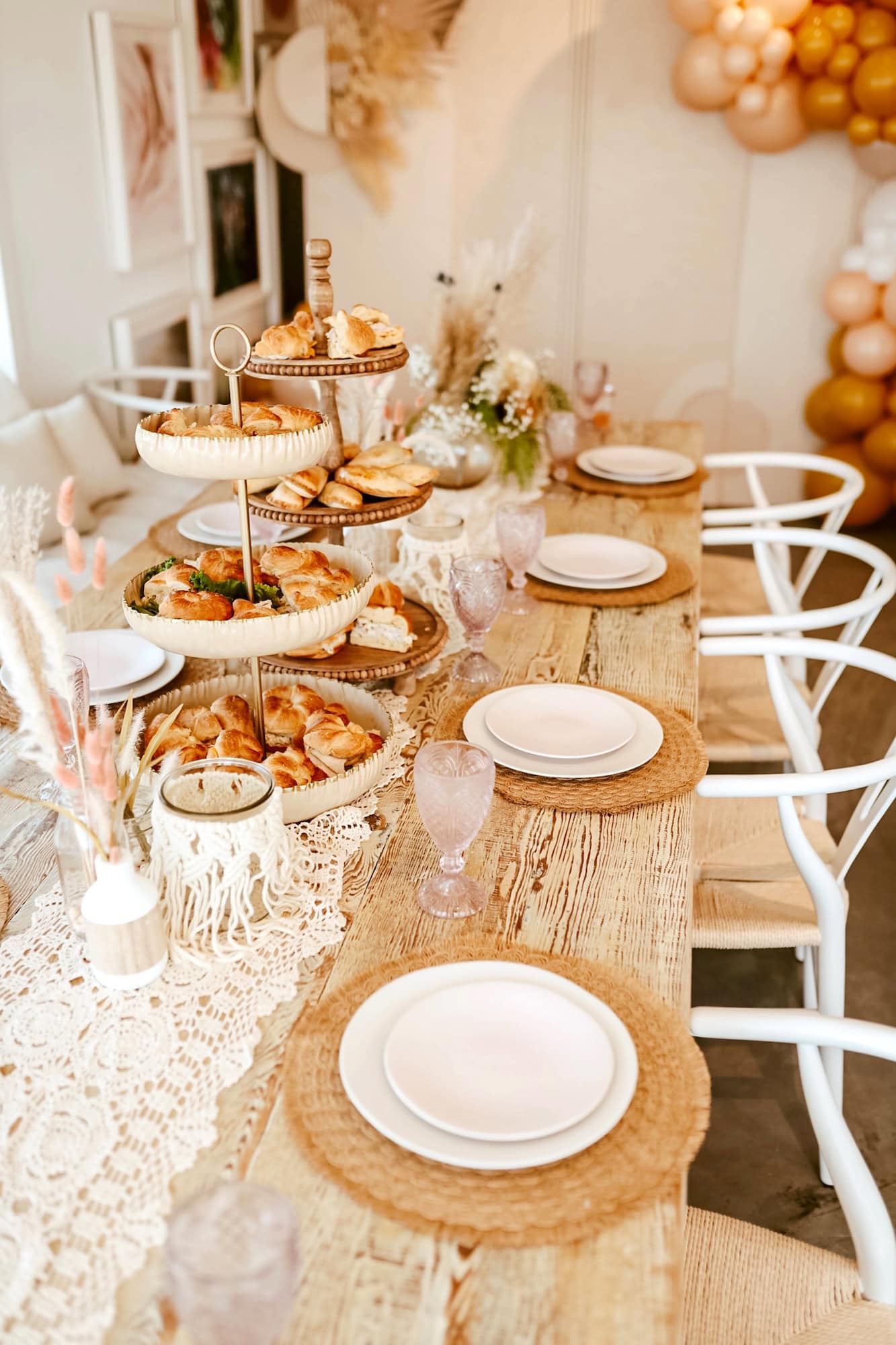 A rustic wooden table is set for a meal with white plates on woven placemats. At the center, a three-tiered stand holds assorted pastries. The boho baby shower decor includes lace, dried flowers, light pink drinking glasses, and in the background are balloons and framed pictures.
