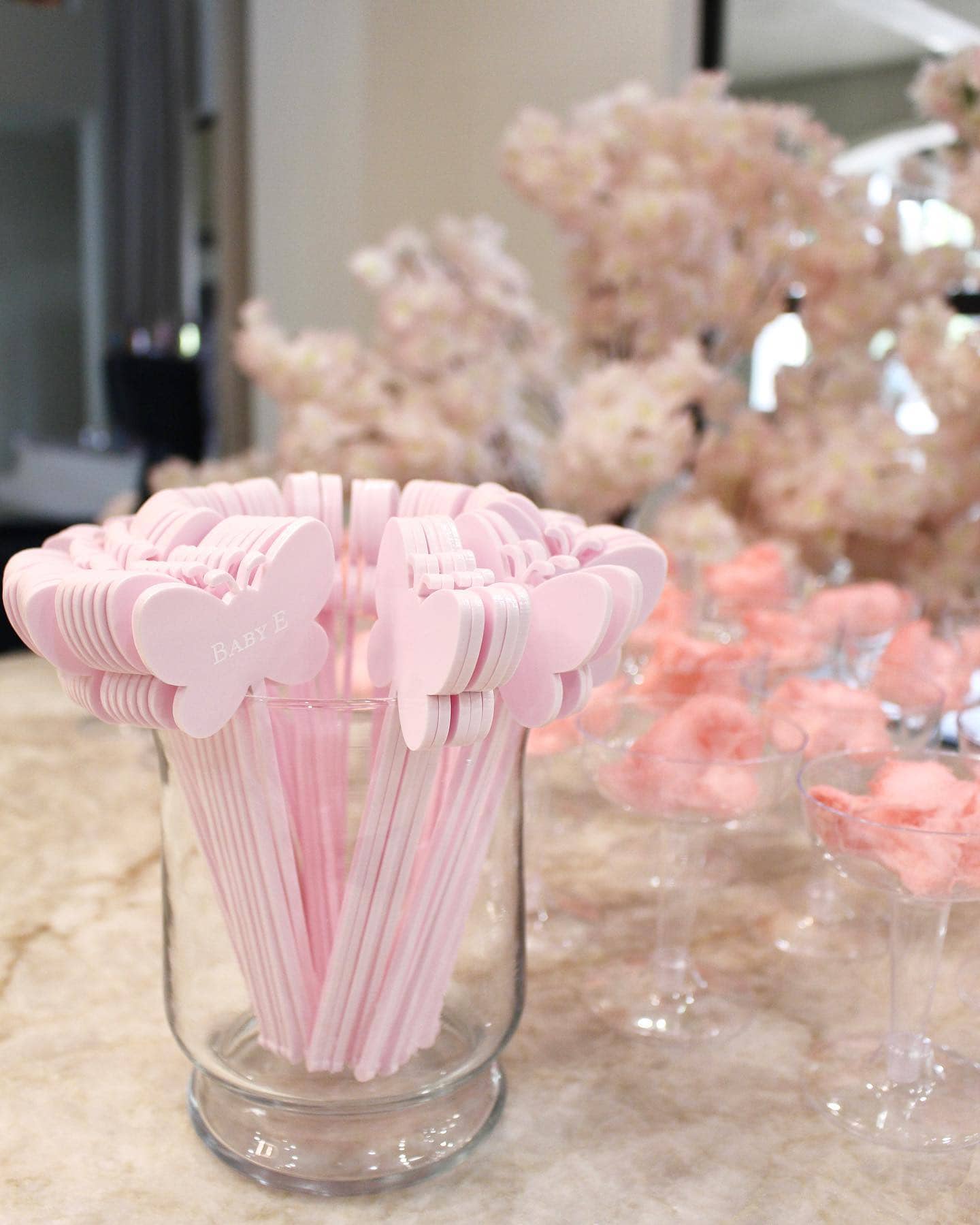 A decorative arrangement for a boho baby shower featuring a glass jar filled with pink butterfly-shaped stirrers labeled "BABY E." In the background, tall glasses of pink cotton candy and delicate pink flowers add to the festive ambiance.