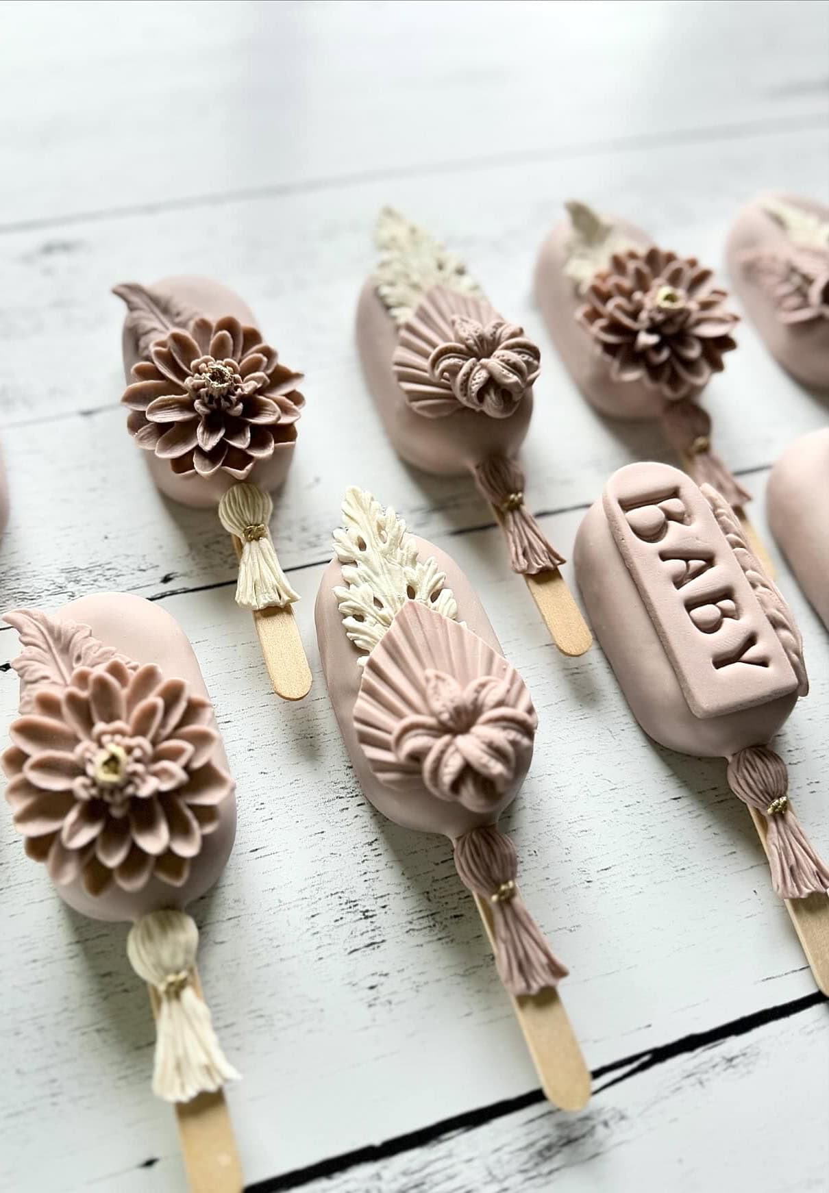 Elegant, blush-toned dessert popsicles are adorned with intricate floral designs, including detailed flowers, leaves, and a bar with the word "BABY" embossed on it. Perfect for a boho baby shower, each treat is beautifully decorated and laid out on a white wooden surface.
