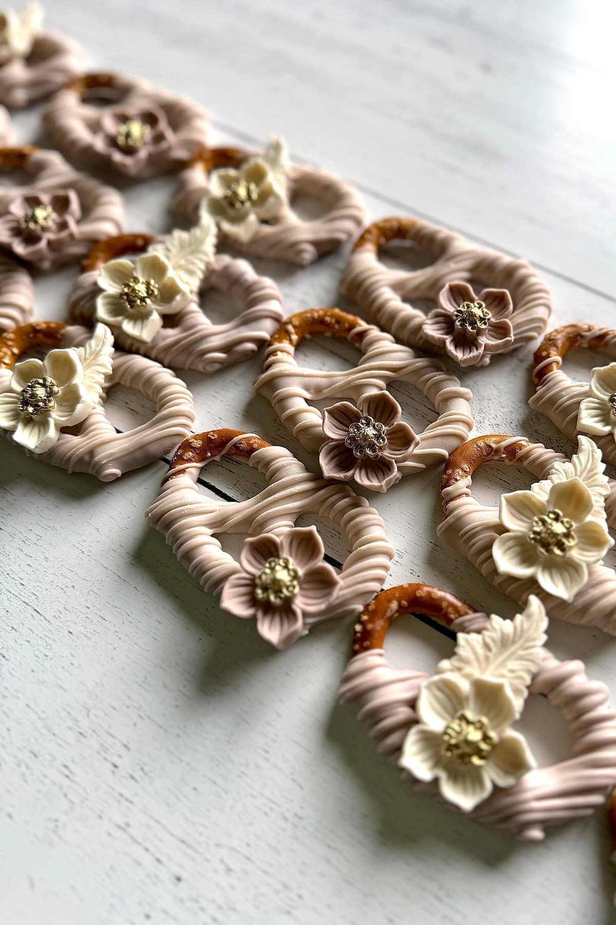 A close-up photograph of beautifully decorated pretzels arranged in neat rows. The pretzels, perfect for a boho baby shower, are adorned with intricate icing in soft beige and cream colors, featuring piped details and delicate flower motifs with pearl-like centers on a white wooden surface.