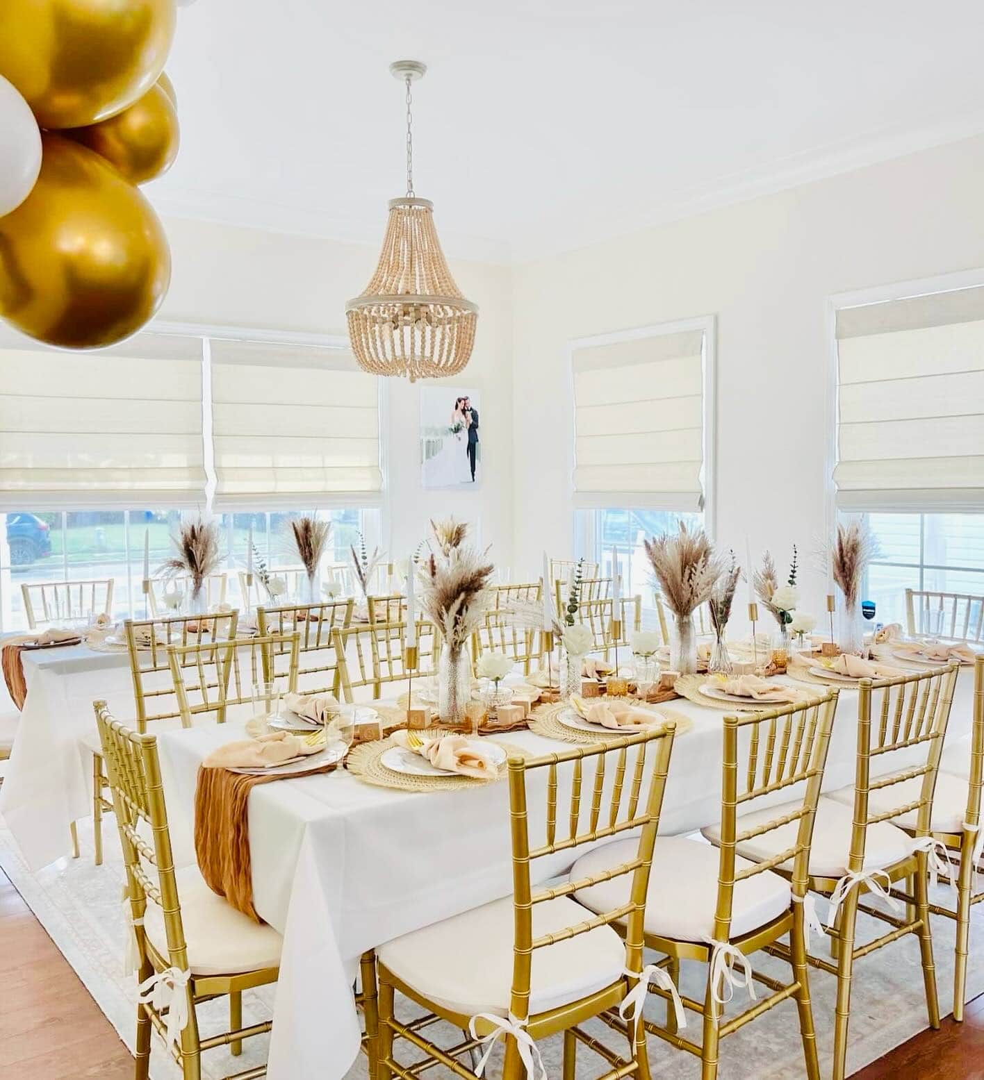 A well-decorated dining room hosts a boho baby shower with two long tables set for a meal. The tables, draped in white tablecloths and surrounded by gold chairs, feature pampas grass centerpieces and beige napkins. A chandelier hangs above, balloons cluster in the upper left corner, and soft natural light fills the room.