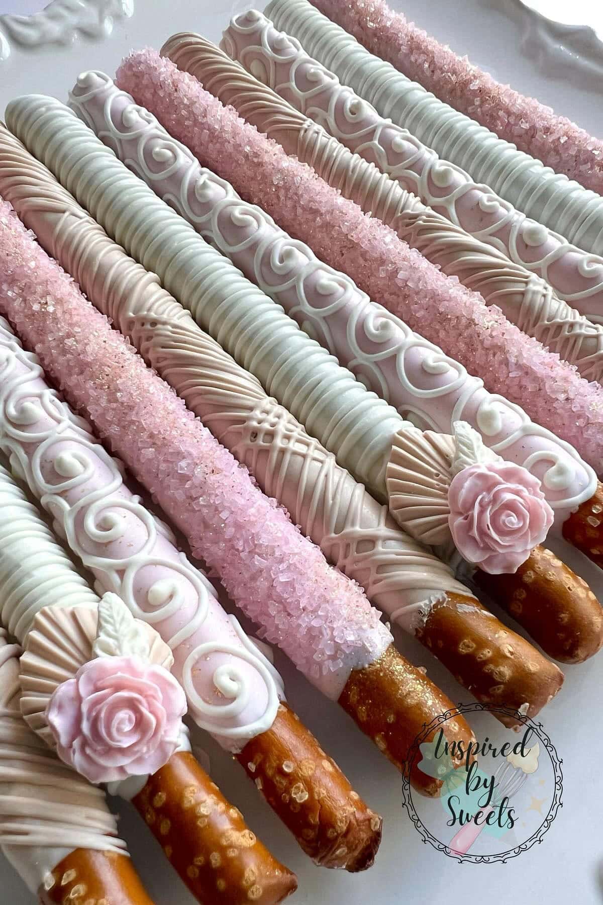 Rows of decorated pretzel rods dipped in white chocolate, embellished with intricate white swirls, pink sugar crystals, and pink rose detailing—perfect for a Boho baby shower. A small round logo in the bottom right corner reads "Inspired by Sweets.