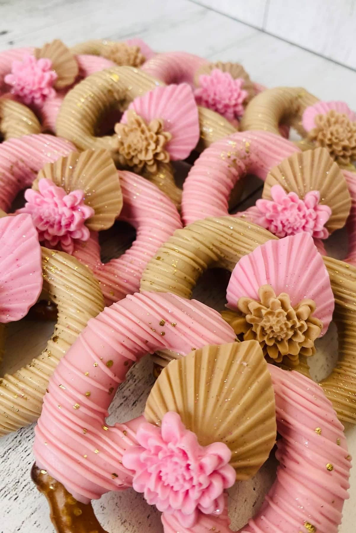 A close-up of decorated pretzels perfect for a boho baby shower. The pretzels are dipped in pink and gold chocolate and embellished with detailed, flower-shaped decorations and striped patterns. Gold glitter is sprinkled on top, adding a touch of sparkle to the treats.