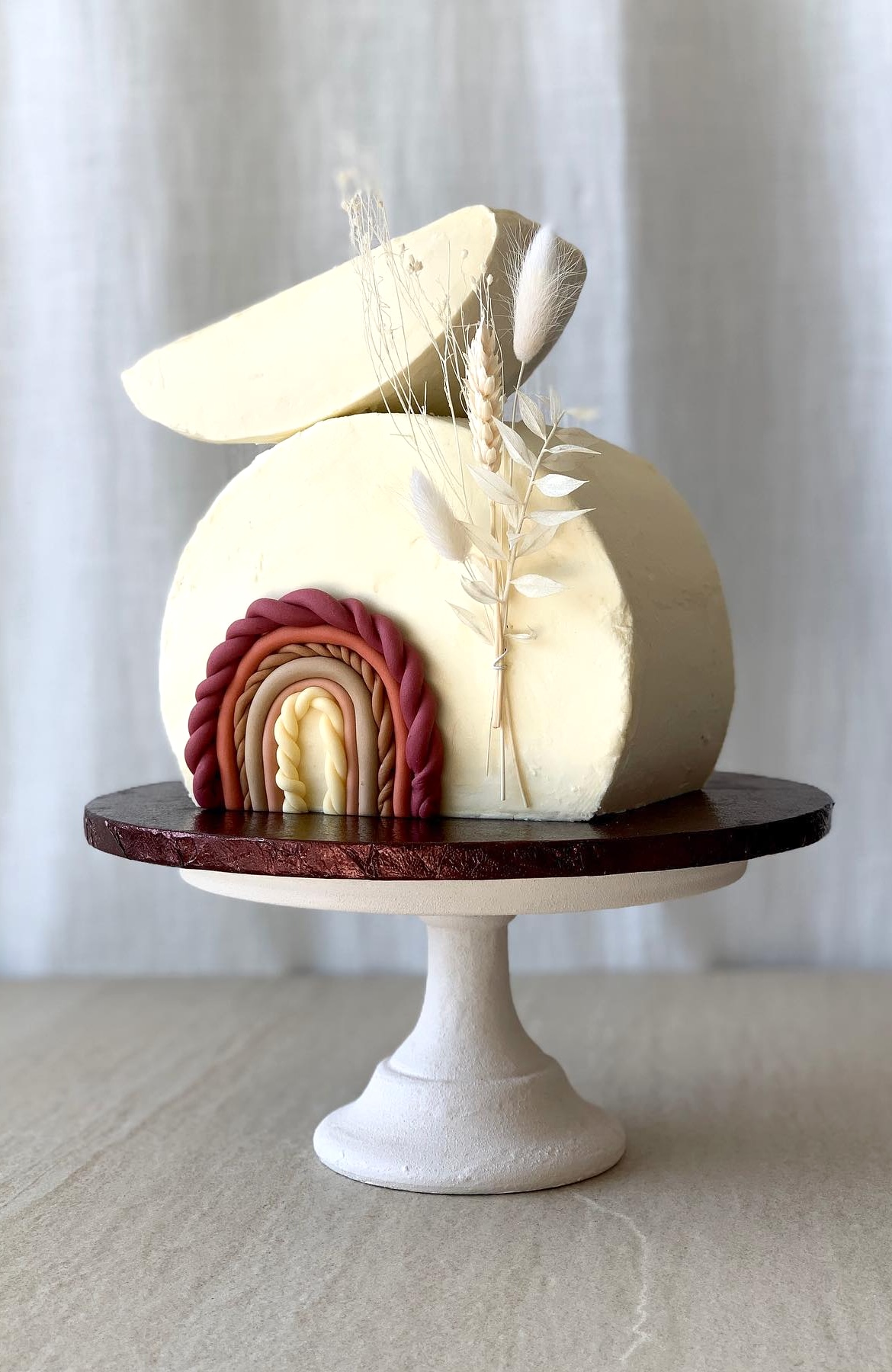 A round, white-frosted cake on a white pedestal stand. Perfect for a boho baby shower, the cake features a brown and beige rainbow decoration and a collection of dried foliage, including wheat, on the side. The background is a light-colored fabric.
