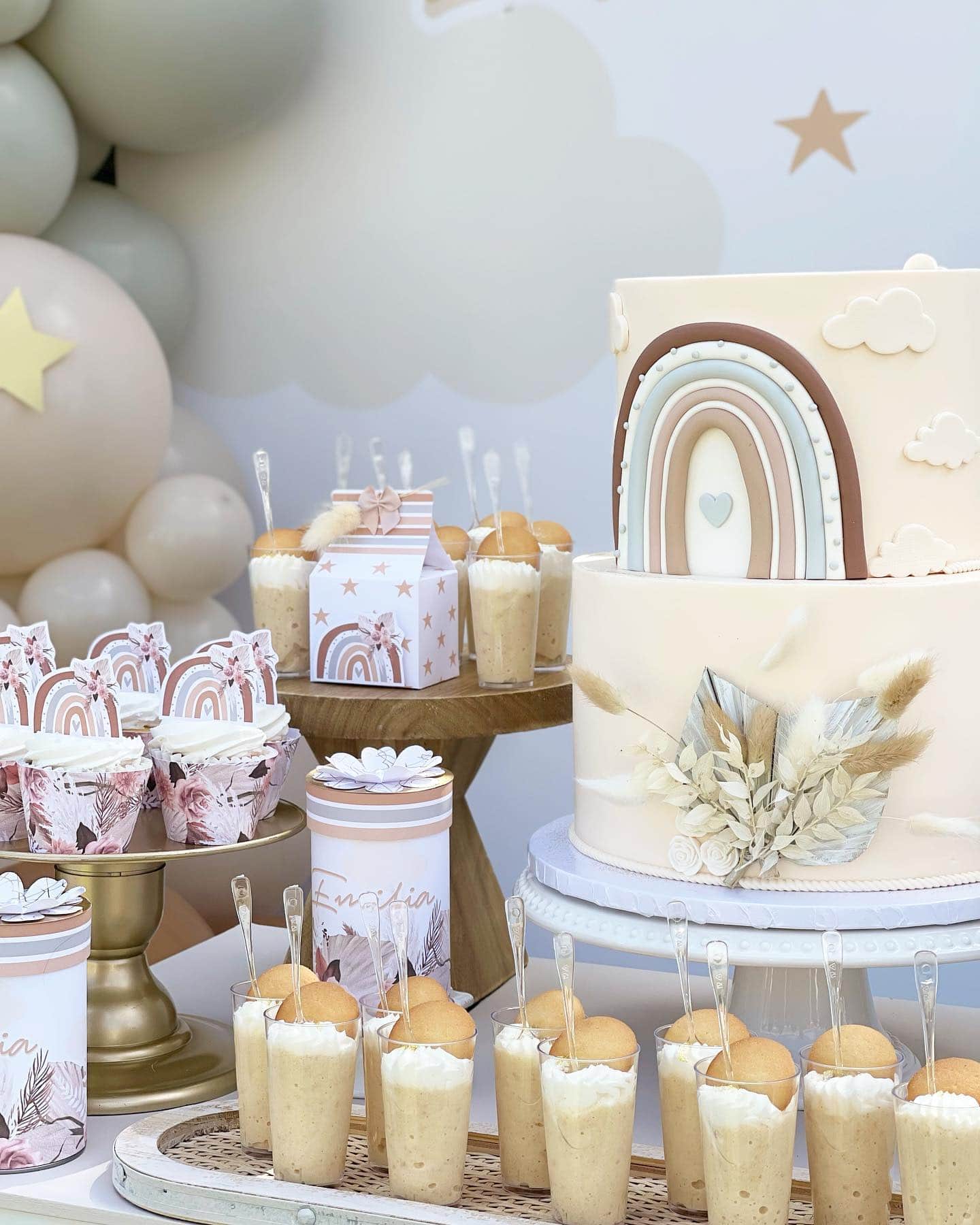 A whimsical dessert table perfect for a boho baby shower, featuring a two-tiered cake with a rainbow and cloud design, small dessert cups with vanilla wafer toppings, decorated cupcakes in pink wrappers, star-shaped paper milk cartons, and two canisters with floral accents. Balloons in the background.