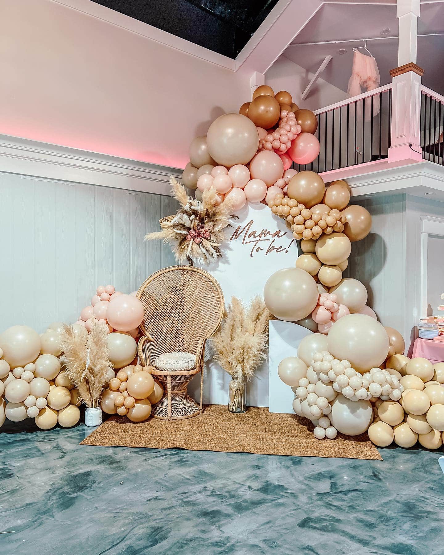 A boho baby shower backdrop features a balloon arch in shades of beige, cream, and pink framing a wicker chair. Pampas grass accentuates the setup, creating a cozy atmosphere, while a staircase and upper level are visible in the background.