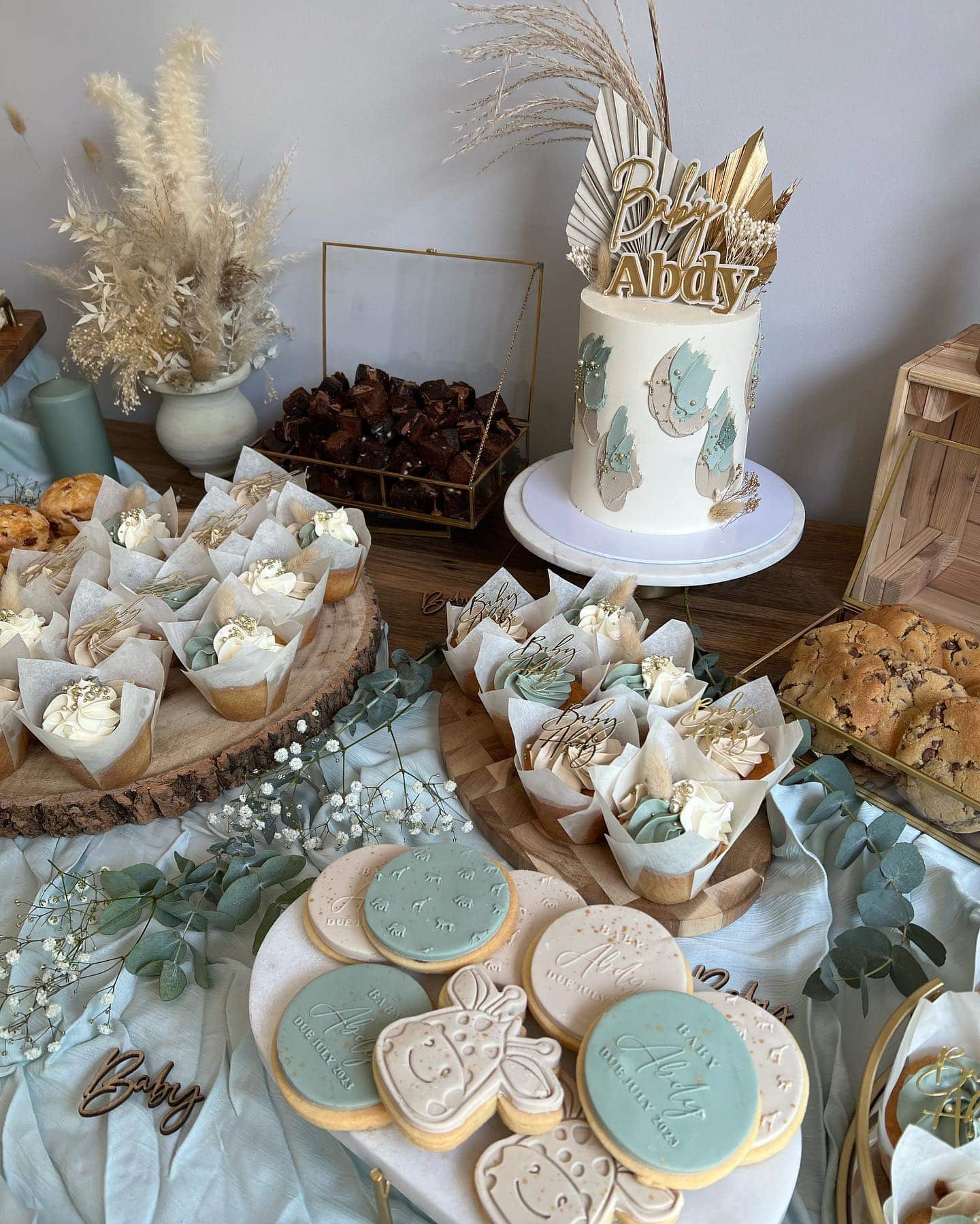 A spread of boho baby shower desserts is displayed on a table. There are iced cookies, cupcakes with floral toppings, a two-tier cake with "Baby" decor, and chocolate brownies. The table is adorned with delicate flowers and elegant decor reflecting a bohemian charm.