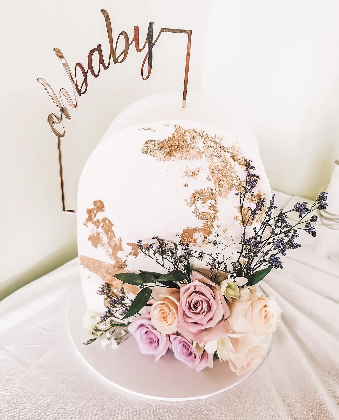 A white cake with gold leaf accents, adorned with a floral arrangement of purple and cream roses, is topped with a decorative sign that reads "ohbaby." Perfect for a boho baby shower, the cake is displayed on a white tablecloth against a neutral background.