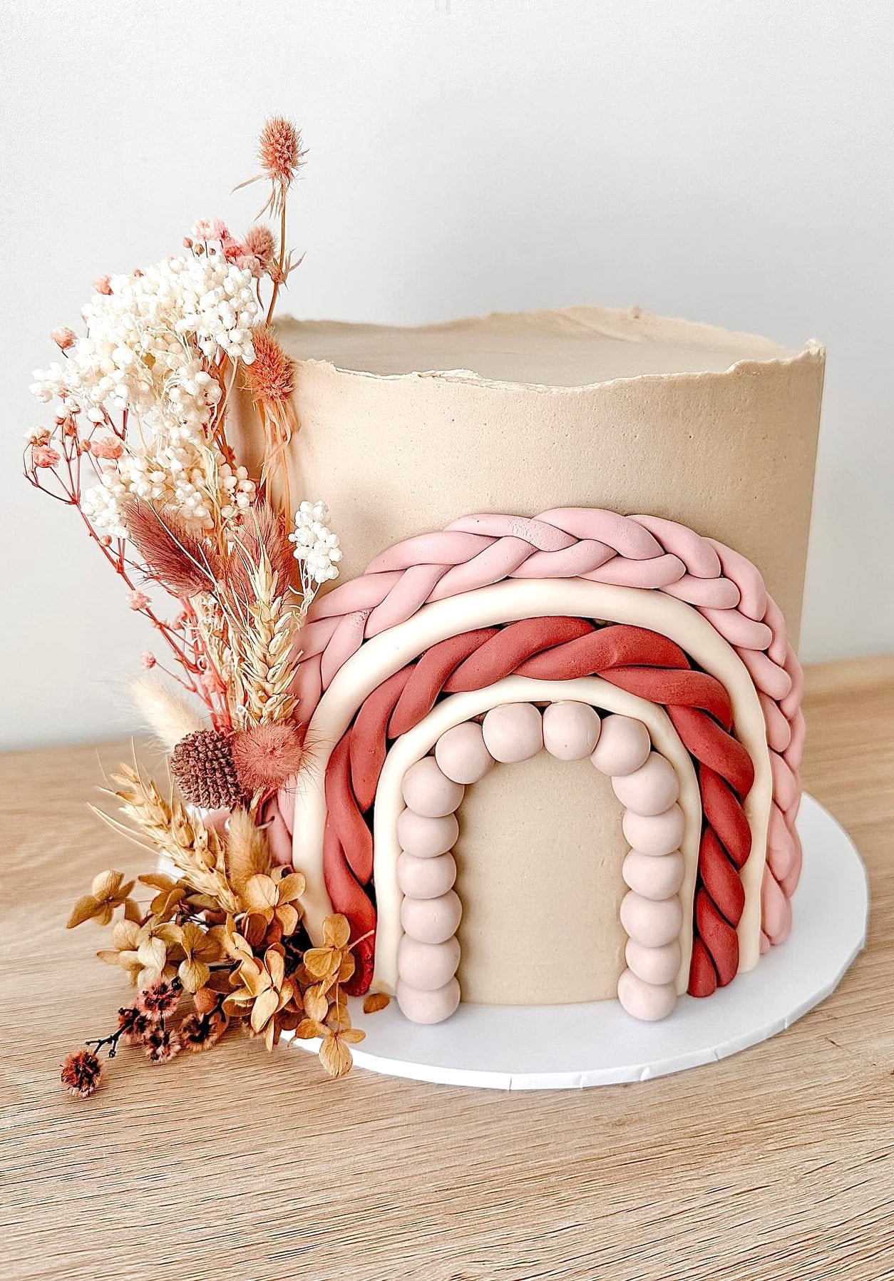A beautifully decorated cake with a smooth beige exterior, adorned with a braided rainbow design in shades of pink and red. Dried flowers and grasses are arranged artistically on one side, creating a rustic and elegant appearance—perfect for a boho baby shower. The cake sits on a wooden table.