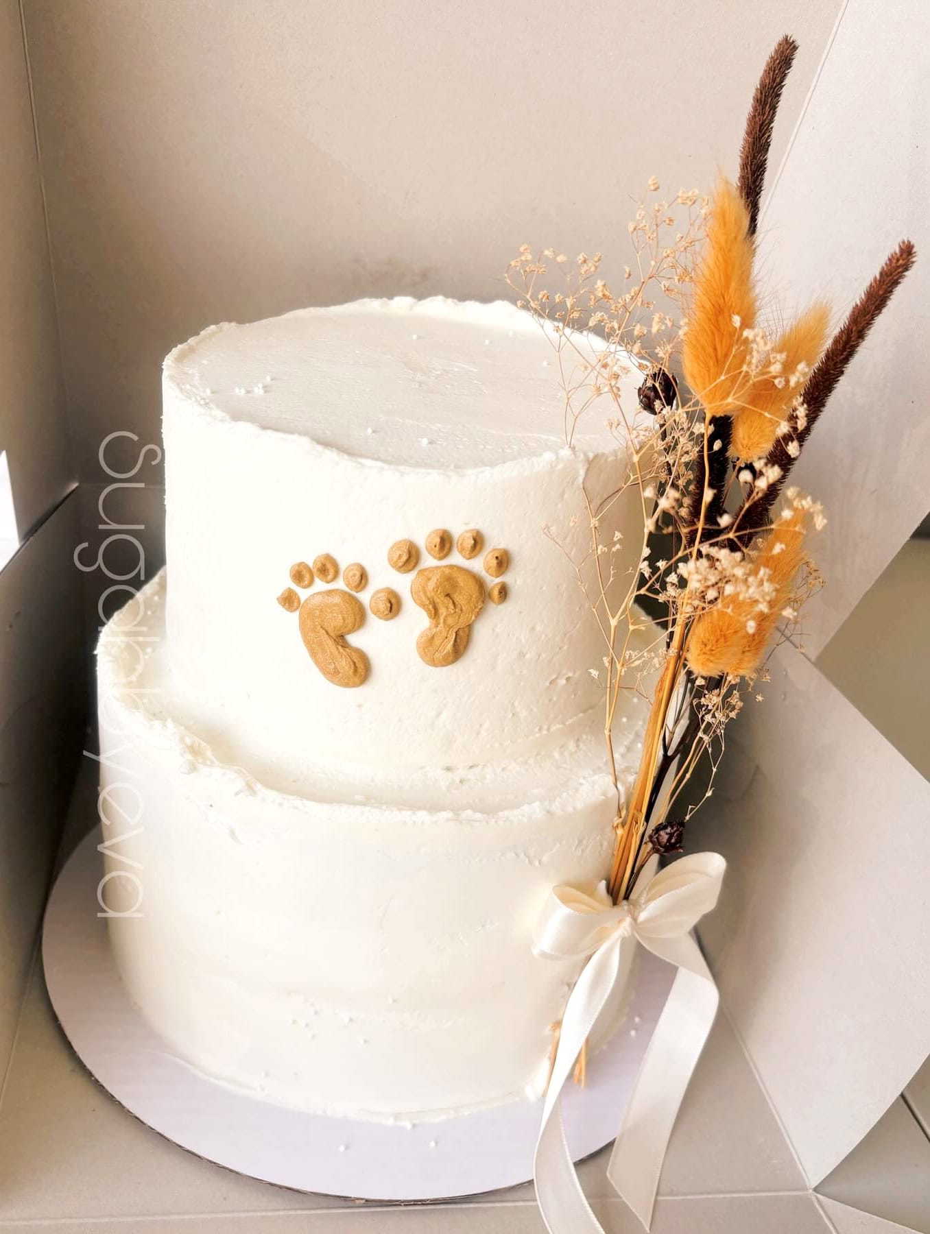 A two-tiered white cake with rustic decorations, perfect for a Boho baby shower. On the upper tier, there are small baby footprints made of cookie dough. The cake is garnished with dried flowers and grasses tied with a white ribbon on the side.