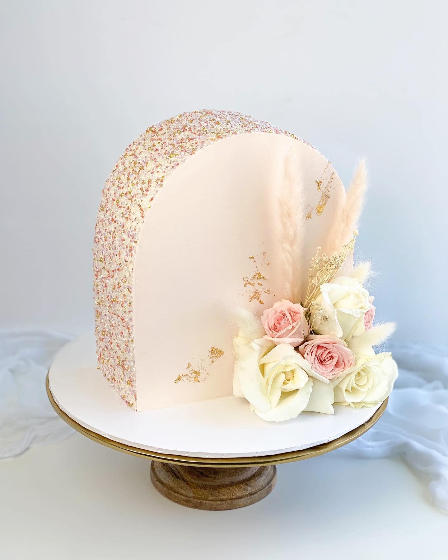 A semi-circular pastel pink cake with a sparkle-covered edge on a wooden cake stand. Adorned with white and pink roses, dried pampas grass, and gold leaf accents, it creates an elegant and delicate look—perfect for a boho baby shower. The background is a plain white setting.