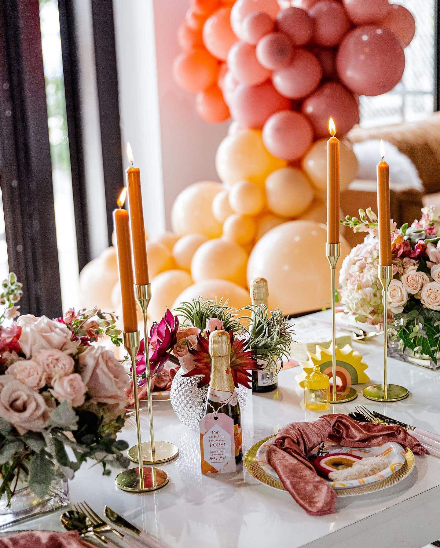 A beautifully set table with a floral centerpiece, long taper candles, and champagne bottles with tags on them. The elegant dishware and cloth napkins enhance the boho baby shower theme. In the background, an eye-catching pink and orange balloon arch completes the festive and inviting setting.