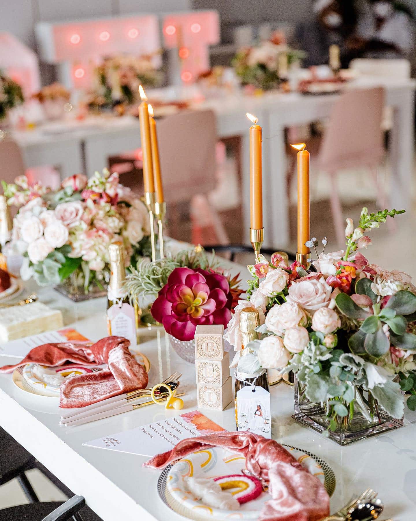 A beautifully set table for a boho baby shower, featuring floral arrangements, lit orange candles, and pink napkins on gold-rimmed plates. Small gifts and place cards adorn the table, with blurred pink and red decor visible in the background.