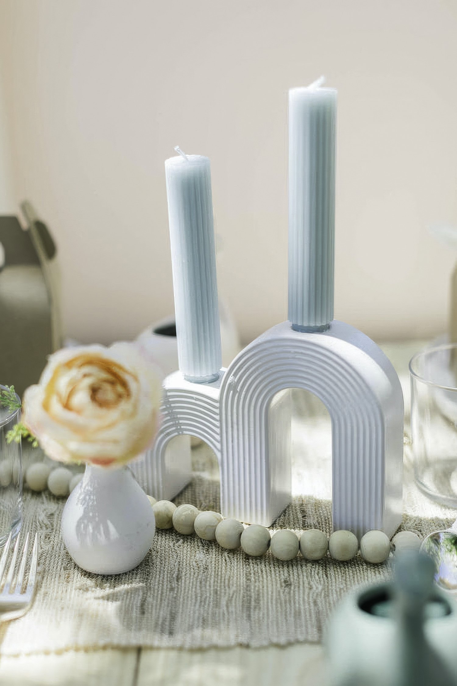 A beautifully set table, perfect for a boho baby shower, features a modern, white, double-arched candle holder with two blue candles. In the foreground, a small vase holds a single peach-colored flower. The table is decorated with a neutral-colored cloth and wooden bead garland.
