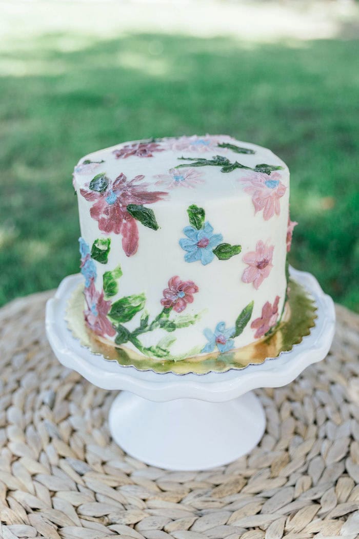 A single-tier white cake adorned with colorful, delicate floral designs in shades of pink, blue, and green sits on a white pedestal cake stand. Perfect for a boho baby shower, the stand is placed on a woven circular placemat with a grassy outdoor background slightly out of focus.
