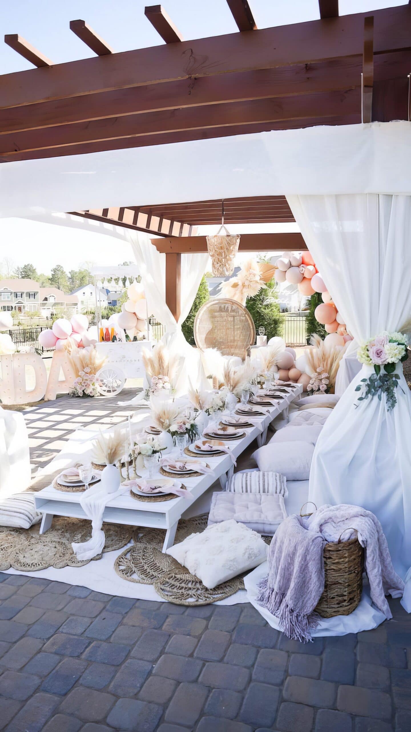 A beautifully decorated outdoor dining space under a wooden pergola, perfect for a boho baby shower. White low tables are adorned with elegant table settings, dried floral arrangements, and pink and white cushions. The backdrop features balloon garlands and draped white curtains.
