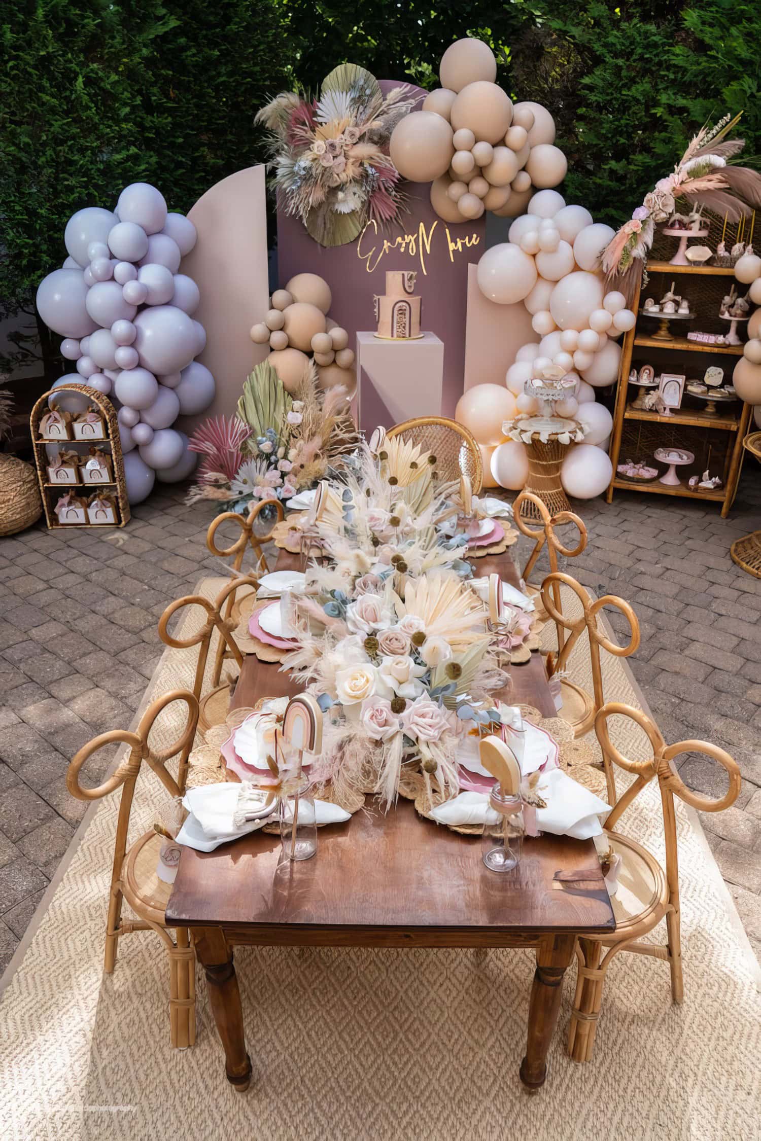 A rustic outdoor dining setup, perfect for a boho baby shower, adorned with neutral-toned pampas grass, white and beige balloons, and elegant floral arrangements. The wooden table is set with white plates, gold cutlery, and dried flowers. A background sign reads "Enjoy Now," creating a cozy ambiance.