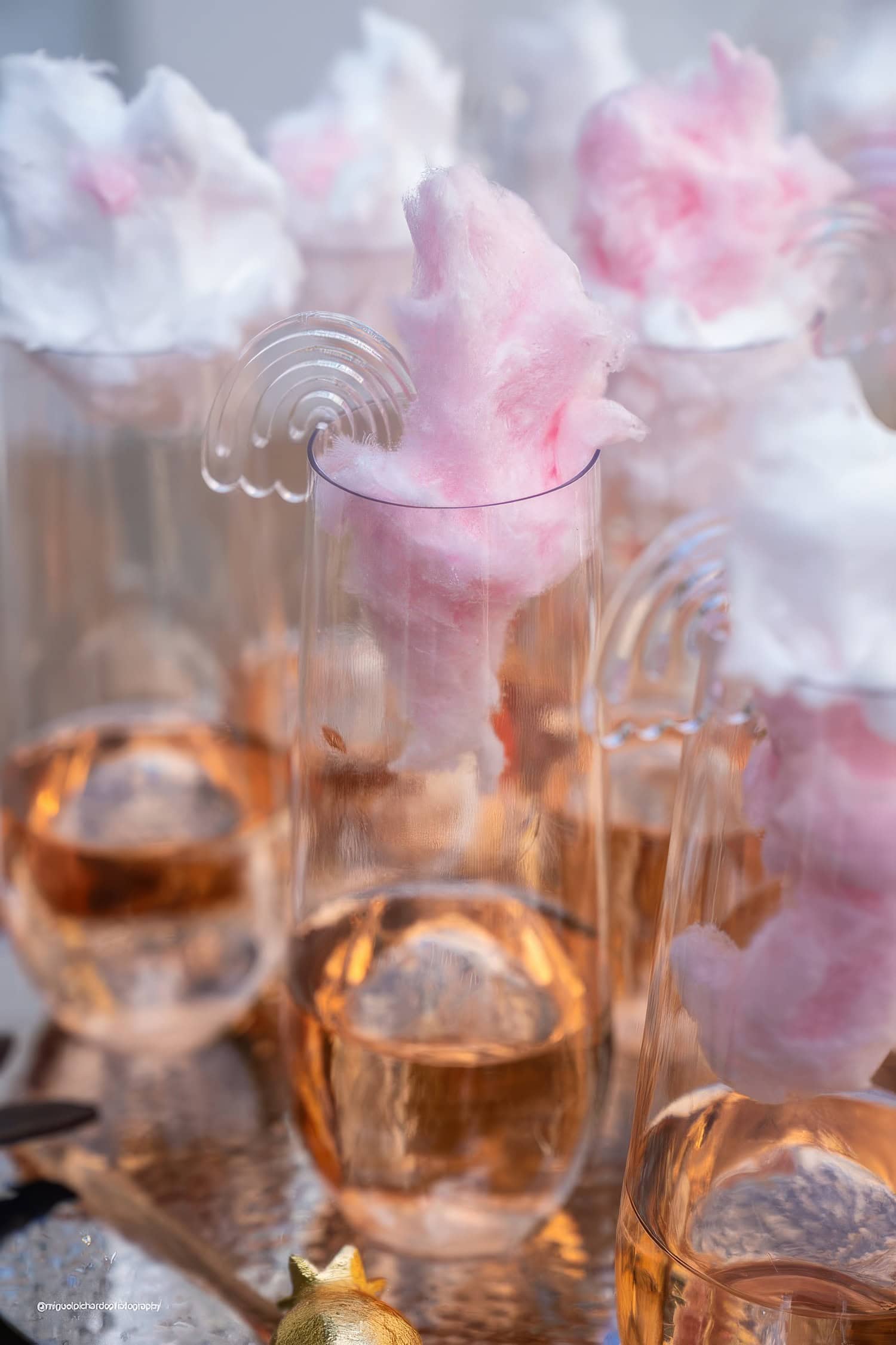 A group of champagne flutes filled with a pinkish beverage are topped with fluffy pink and white cotton candy. The glasses, perfect for a boho baby shower, are arranged on a reflective surface, creating an elegant and whimsical aesthetic.