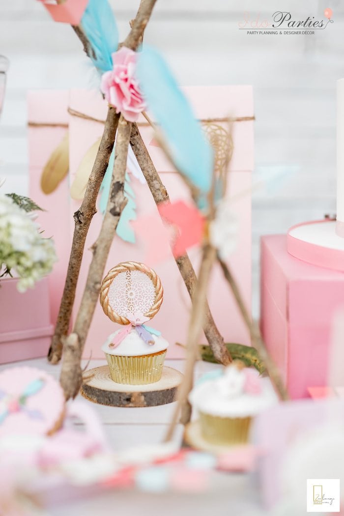 A pastel-themed party setup perfect for a Boho baby shower features a cupcake topped with a dreamcatcher decoration. The cupcake is placed under a small teepee made of twigs, surrounded by soft pink and blue accents, flowers, and other decorative items.