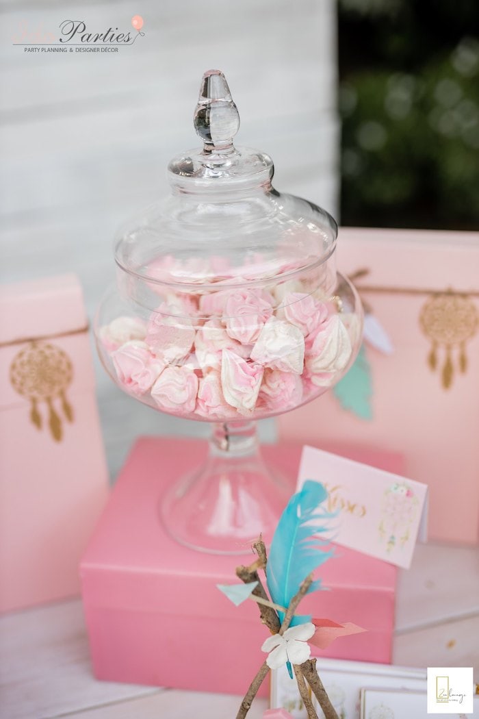 A clear glass apothecary jar with a lid is filled with pink and white meringues. The jar sits on pink boxes adorned with dreamcatcher illustrations and ribbons, perfect for a boho baby shower. Colorful feathers and a greeting card complete the charming decorative setup.