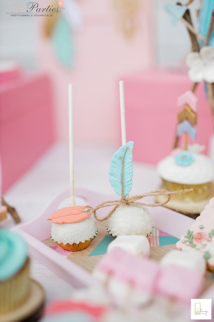 Two cake pops with white coating, decorated with colorful fondant feathers, are placed upright in a pink tray. Surrounding them are various pastel-colored desserts, perfect for a Boho baby shower, including a cupcake with a blue frosting flower and other sweets on a white table.