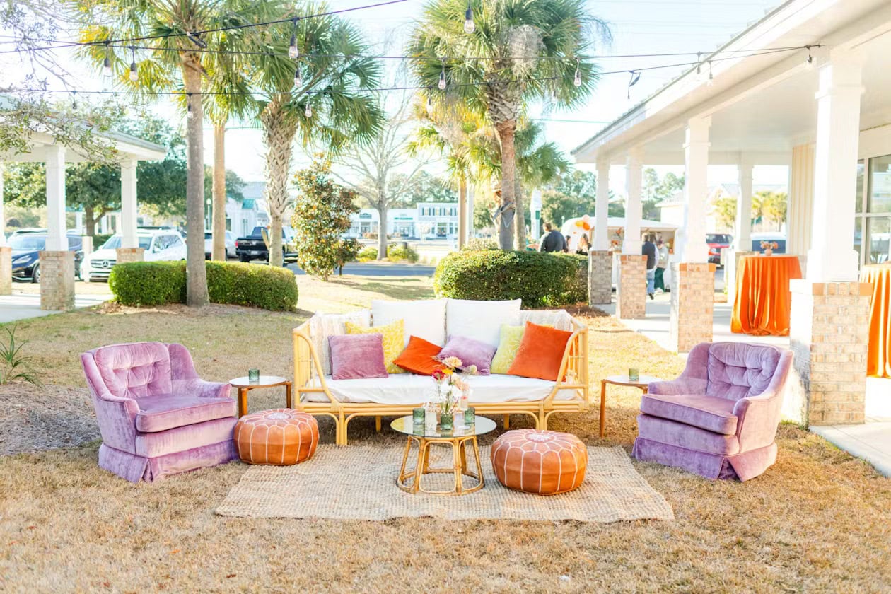 Outdoor seating area with a cozy setup including a white sofa adorned with pillows, two purple armchairs, and two orange ottomans placed on a woven rug. A small round table with flowers is central, creating the perfect boho baby shower vibe. Palm trees, a white building, and cars are in the background.