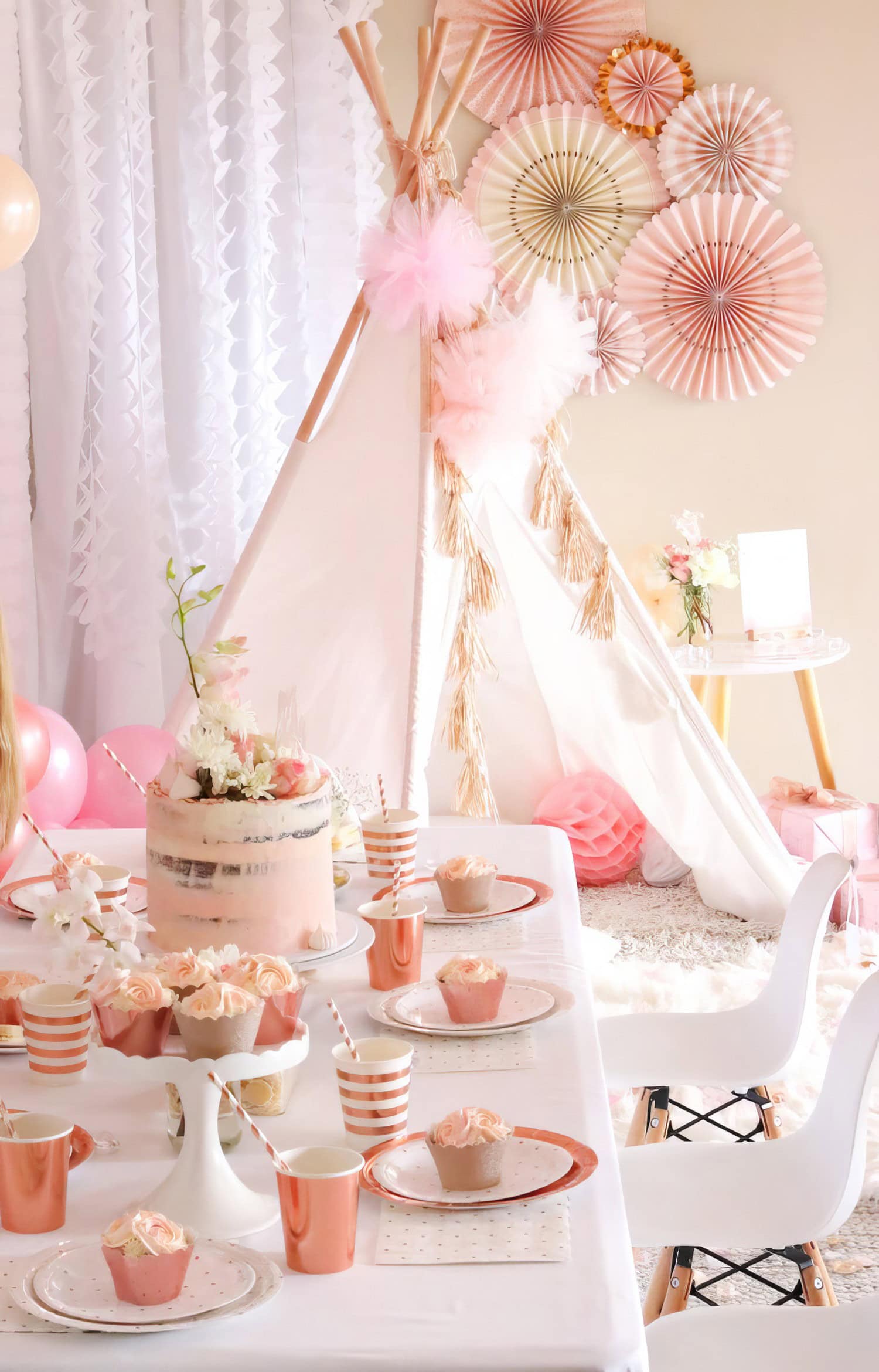 A chic, pastel-themed boho baby shower setup features a white teepee adorned with pink decorations in the background. A table is set with pink plates, rose gold striped cups, cupcakes, and a large pink frosted cake topped with flowers. White chairs surround the table.