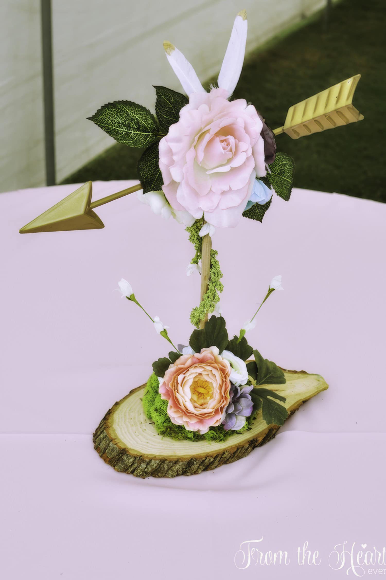 A decorative arrangement featuring a pink rose, green leaves, and arrow accents held in place on a wooden slab sets the tone for a boho baby shower. Placed on a light pink tablecloth with grass in the background, the heartfelt words "From the Heart" are partially visible at the bottom.