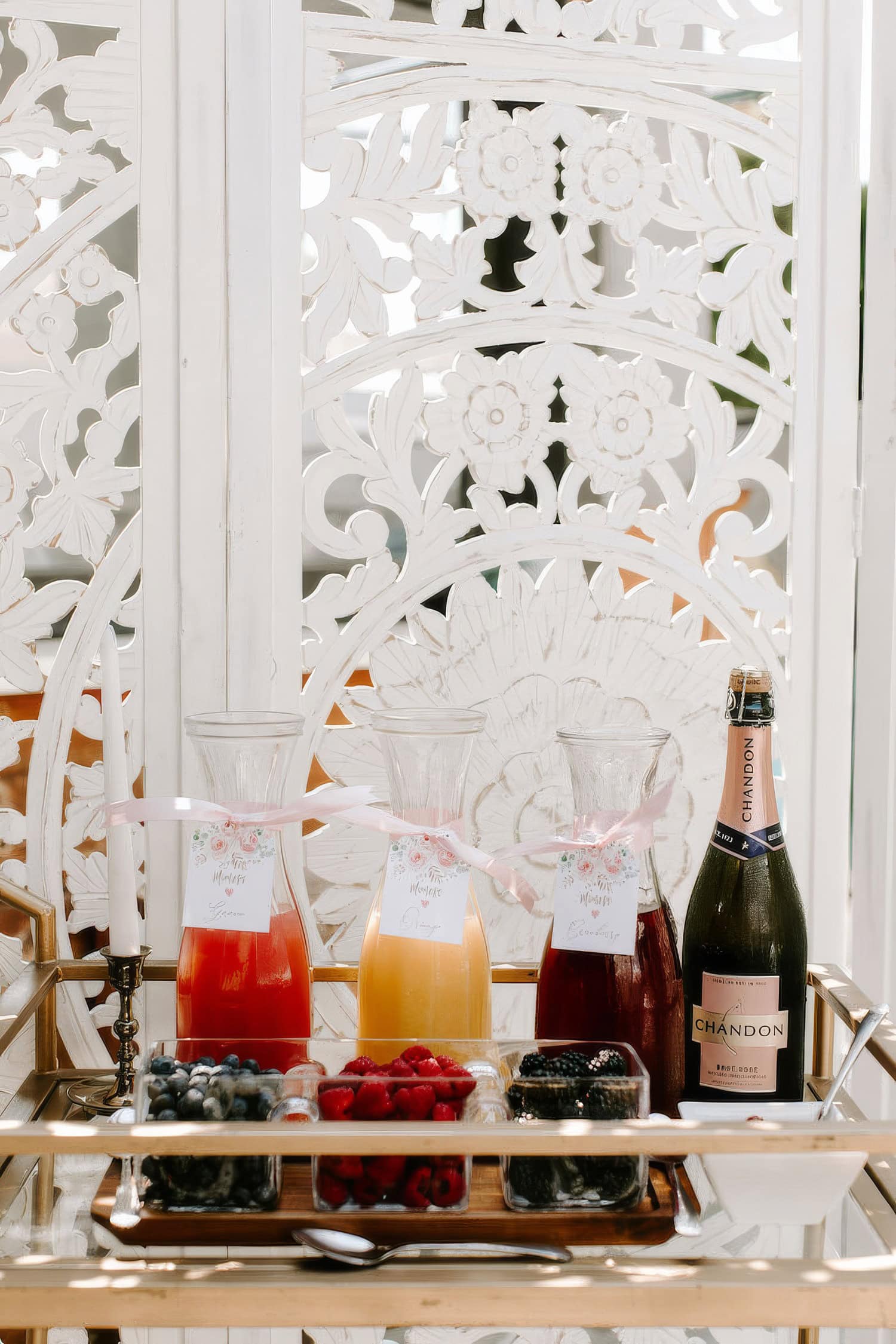 A stylish bar cart holds glass carafes filled with red, orange, and pink juice, each adorned with a ribbon and tag. A bottle of Chandon champagne rests beside the carafes. Small bowls of fresh berries and a candle complete the elegant boho baby shower setup, backed by ornate white panels.